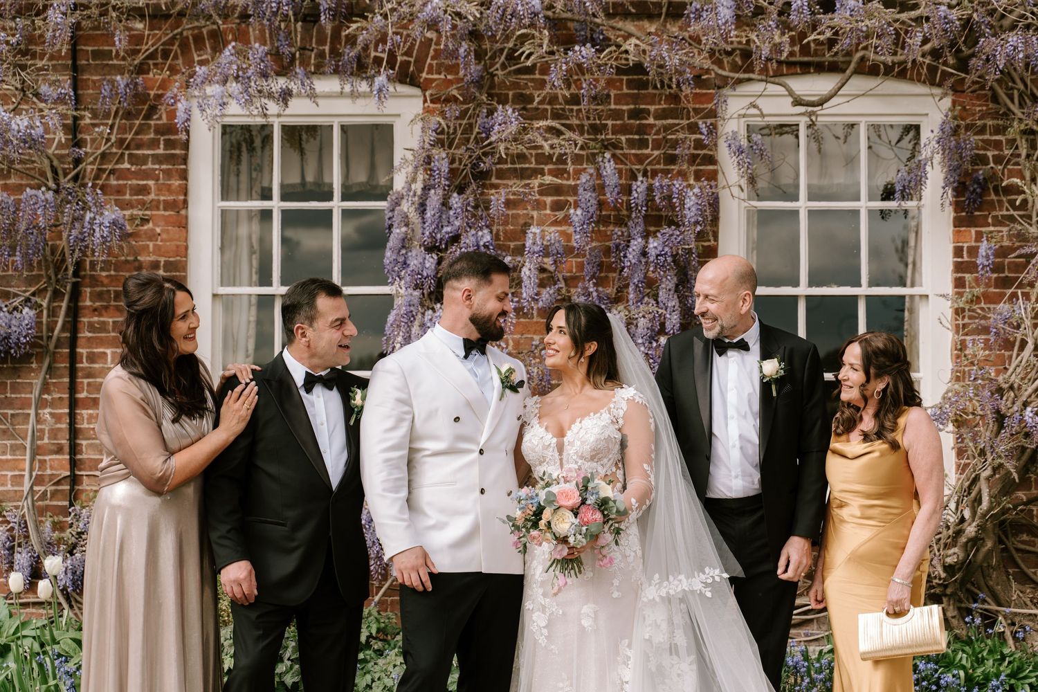 Wedding party poses before brick wall with wisteria, bride in lace gown holds bouquet, groom in white tuxedo jacket.