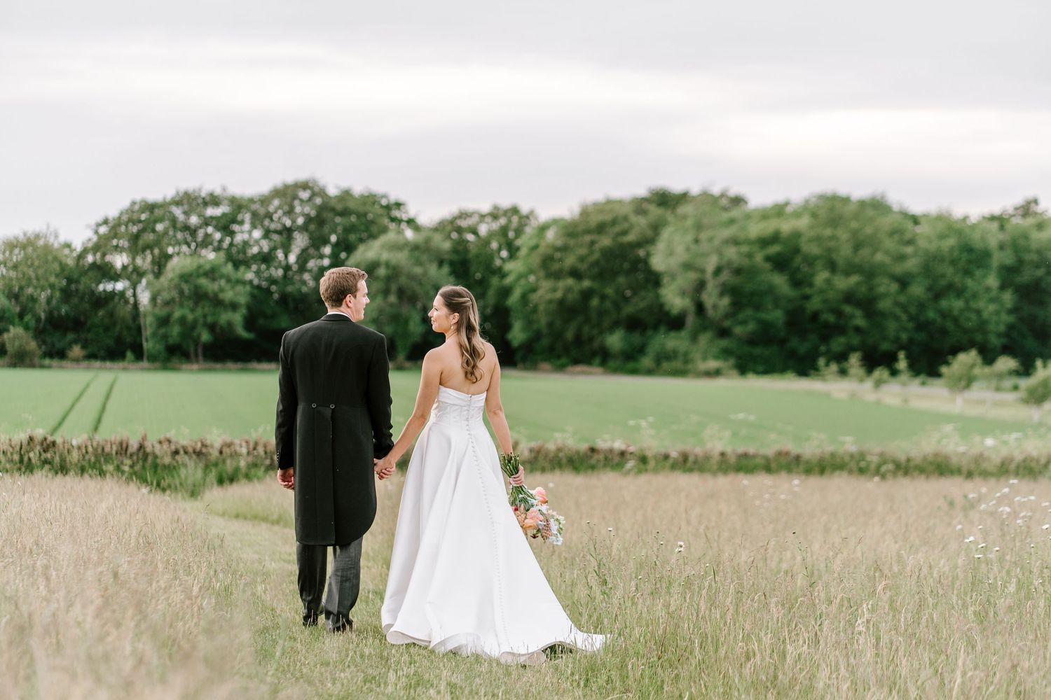 Bride and groom walking hand in hand through a lush green field on their wedding day.