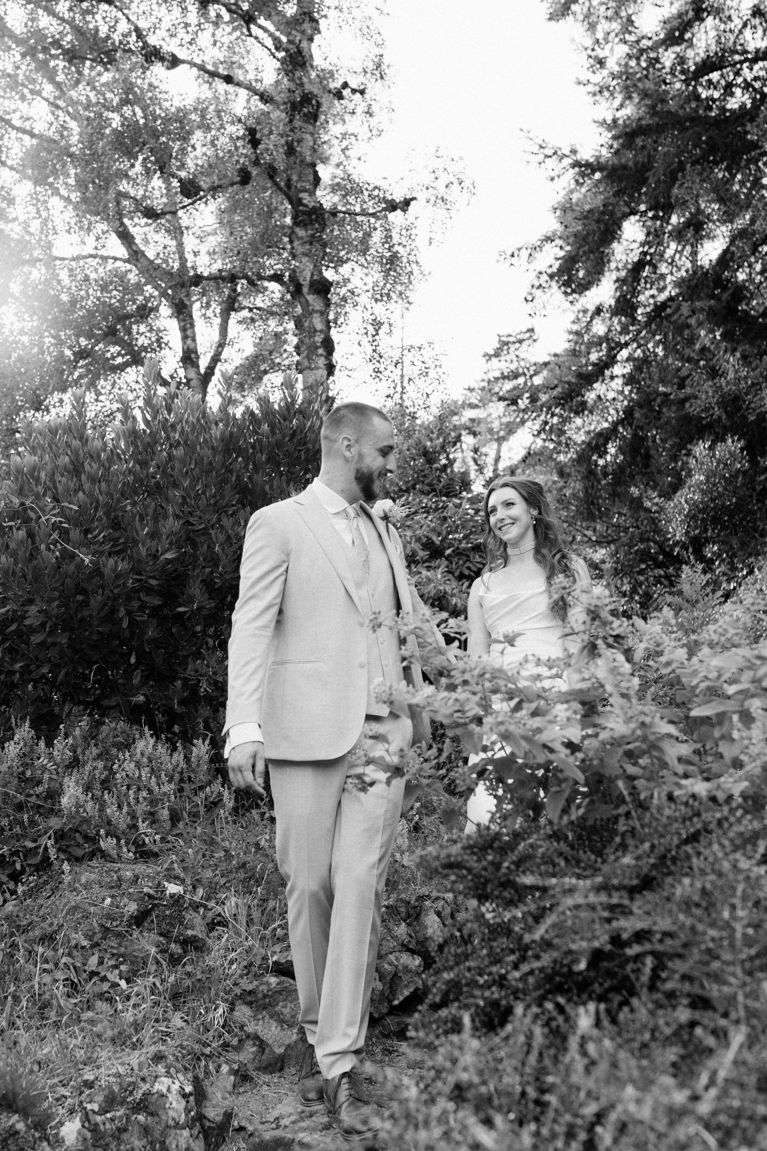 Black and white photo of bride and groom sharing a moment among lush garden greenery in sunlight.