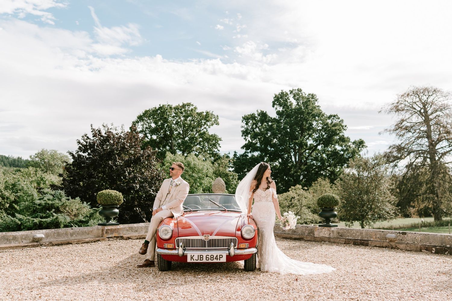 Bride and groom leaning against a vintage red MGB convertible car at an elegant countryside wedding venue.