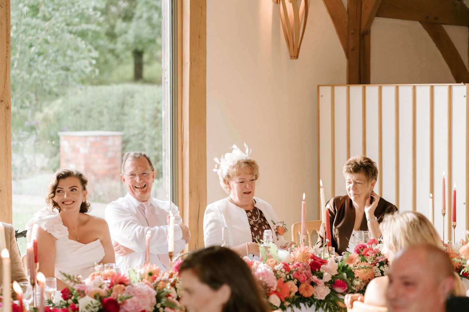 Wedding party seated at head table with colorful floral arrangements, candles, and guests laughing during reception.