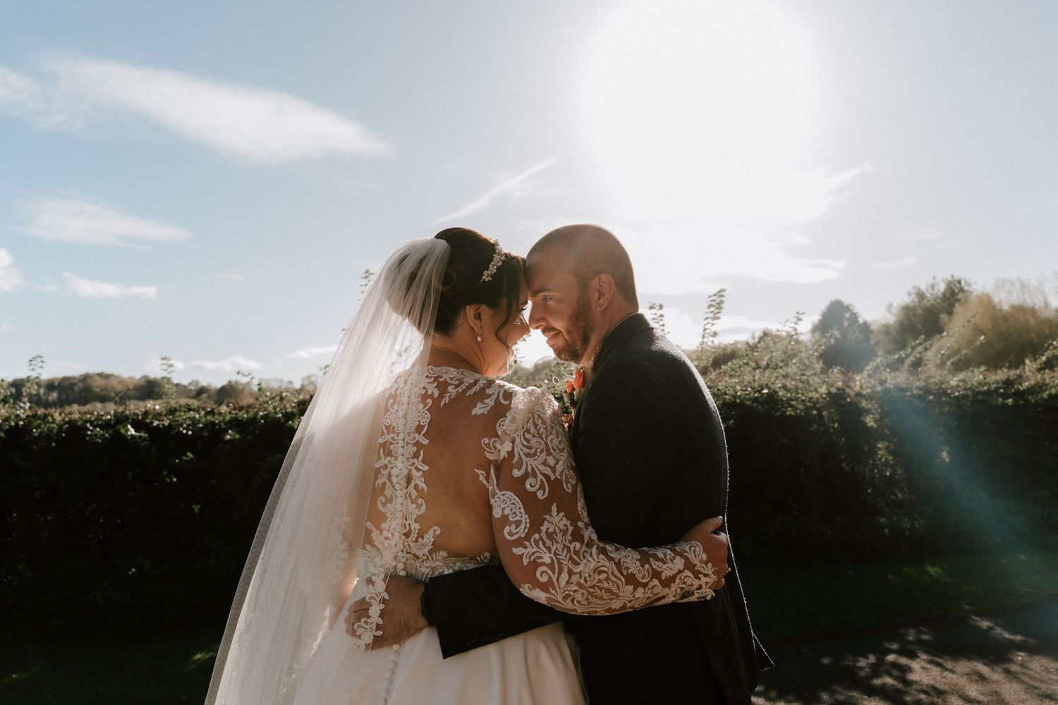 Bride and groom sharing an intimate moment outdoors, backlit by golden sunlight with lush green hedges behind them.