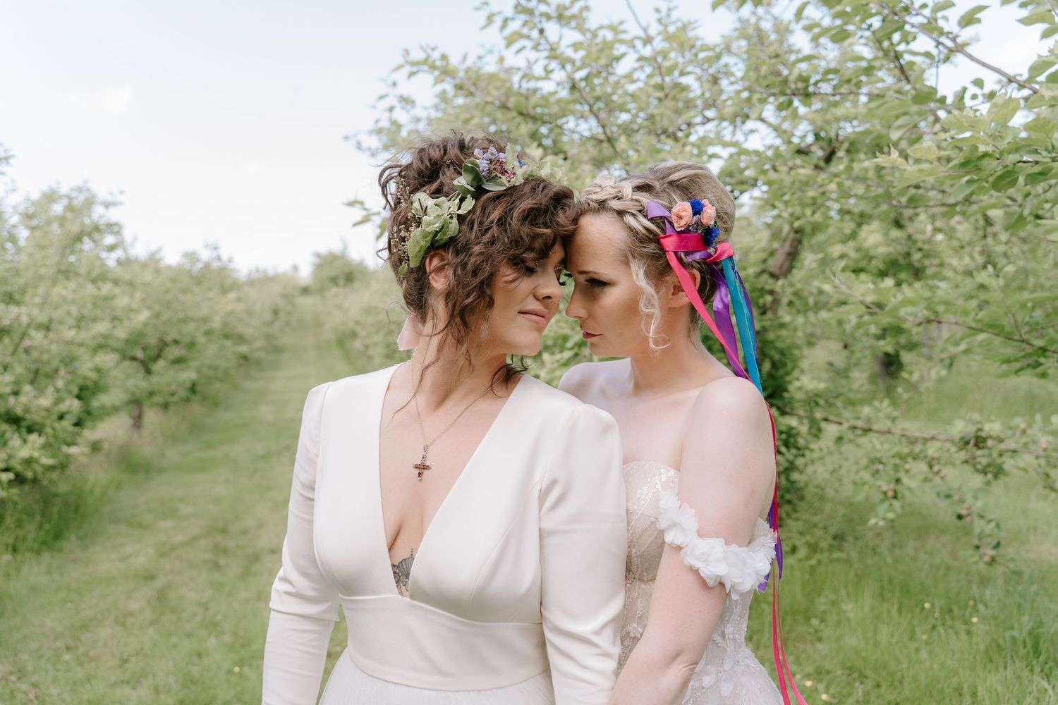 Two brides in a lush green orchard, one in white gown with floral crown, share an intimate moment.