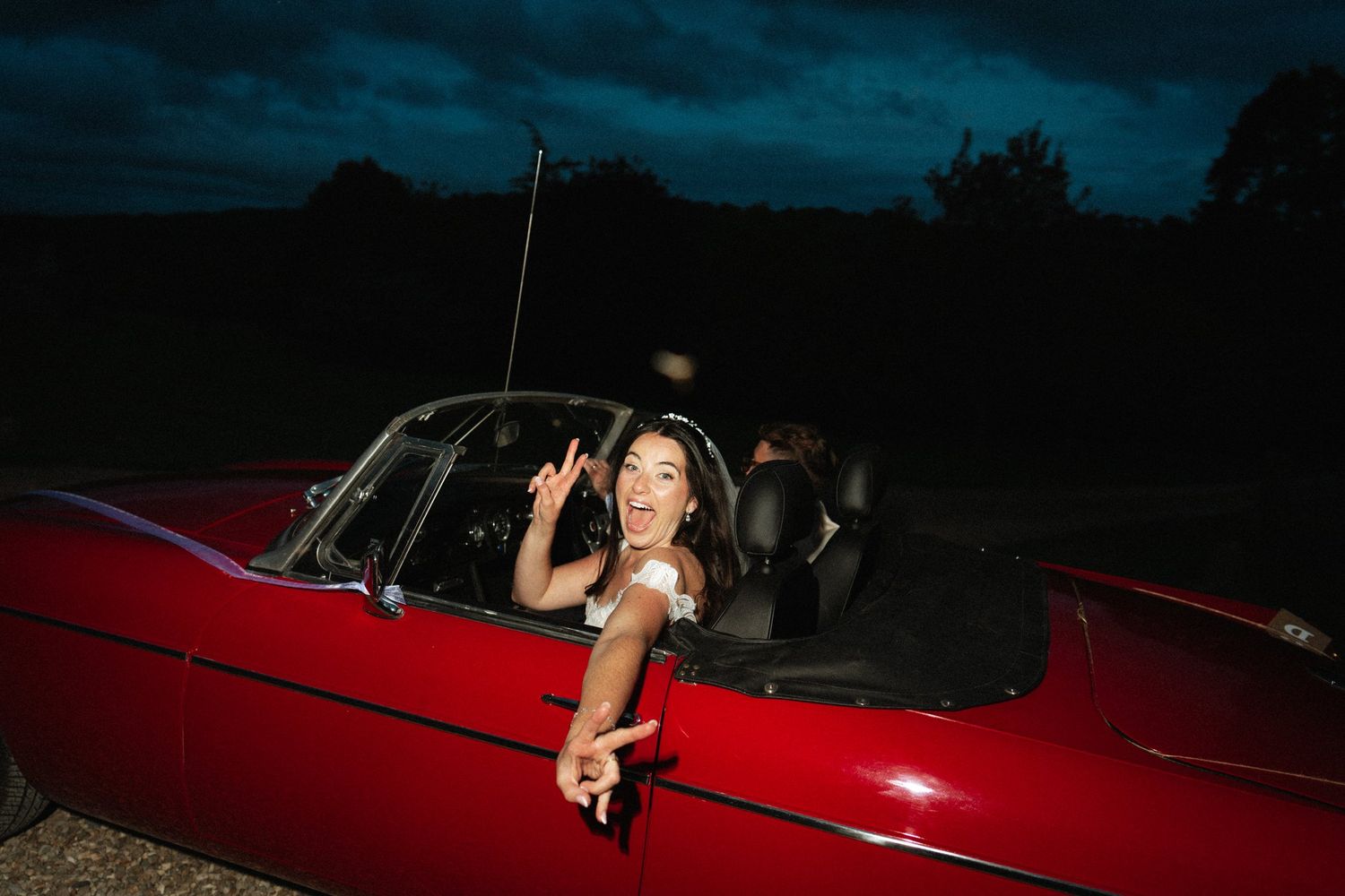 Woman smiling and making peace sign from red convertible classic car at night.