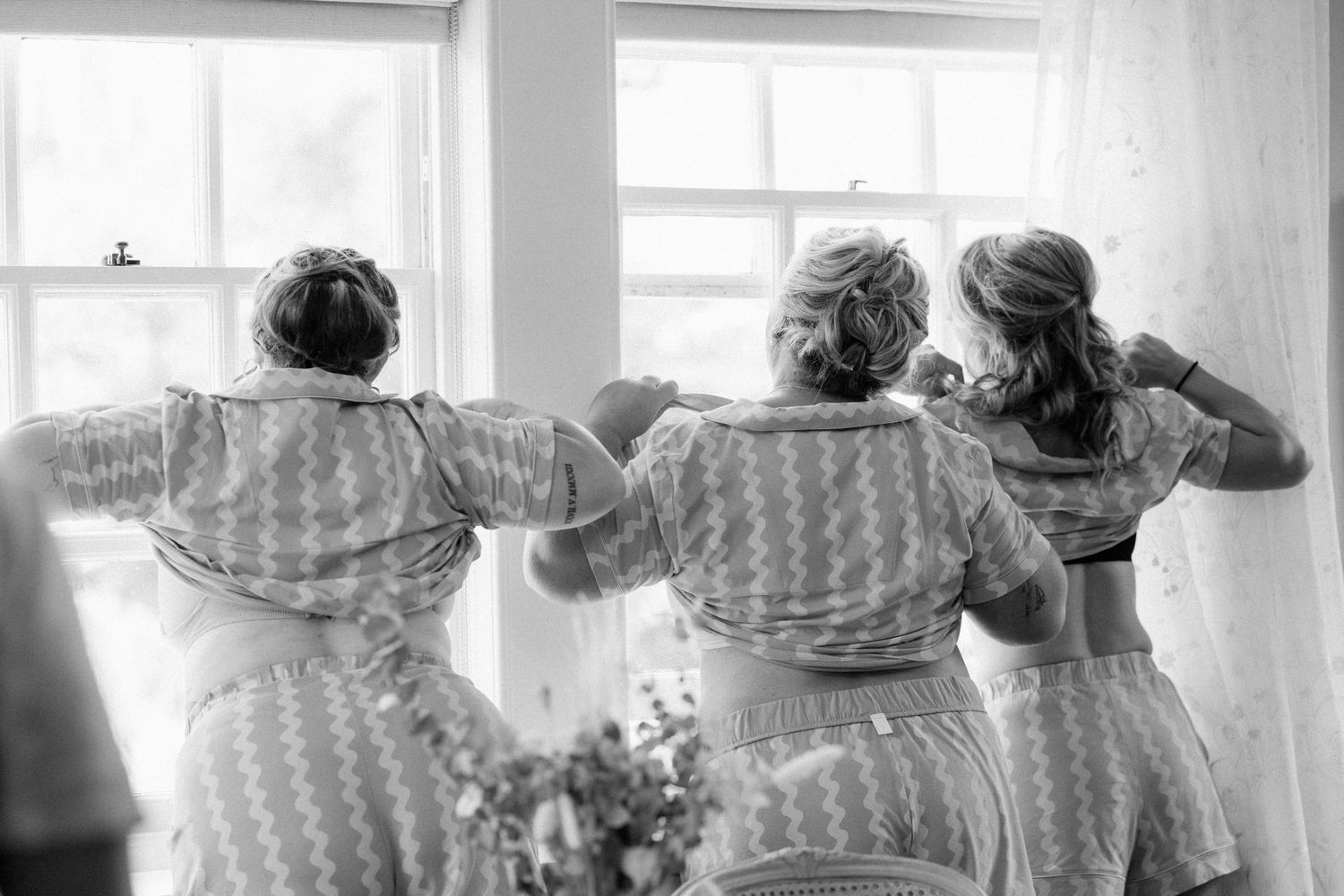 Black and white photo of three bridesmaids in matching dresses standing by a window adjusting their hair before a wedding.