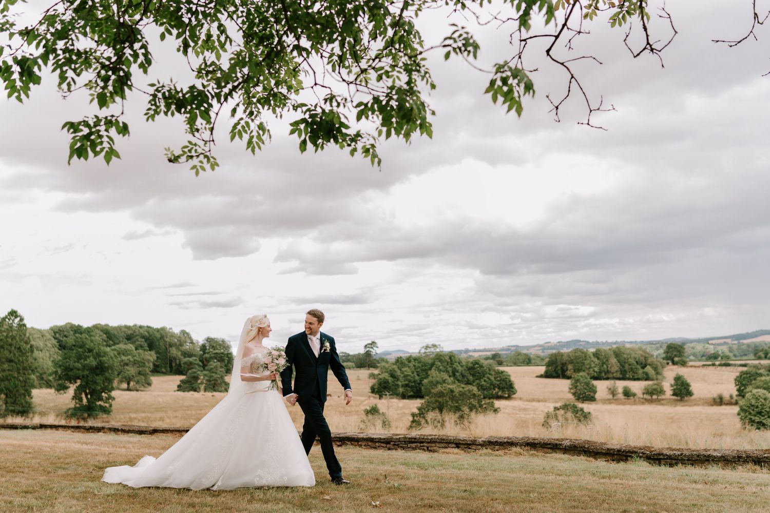 Bride and groom walking hand in hand through golden fields under cloudy skies at a rustic outdoor wedding.