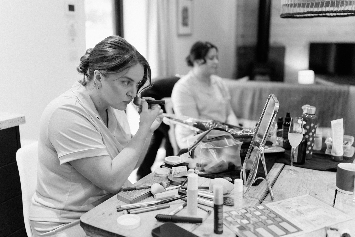 Black and white photo of a woman applying makeup at a vanity table covered with beauty products.