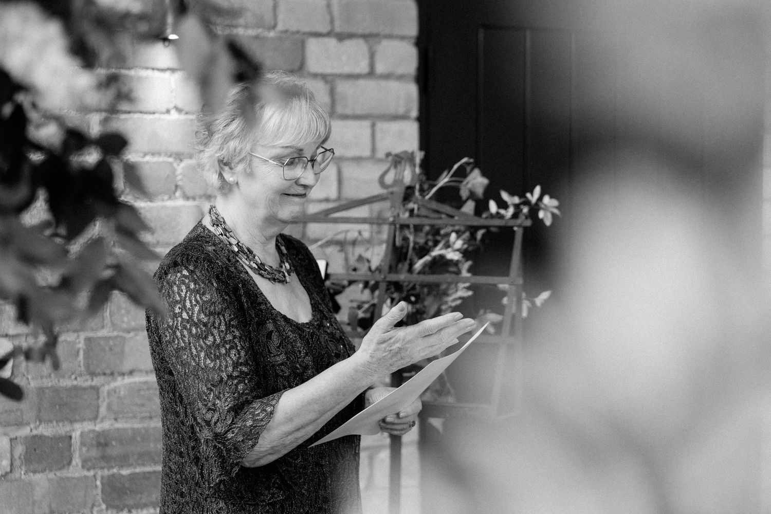 Elderly woman in black lace dress reading from a clipboard during an outdoor wedding ceremony by a brick wall.