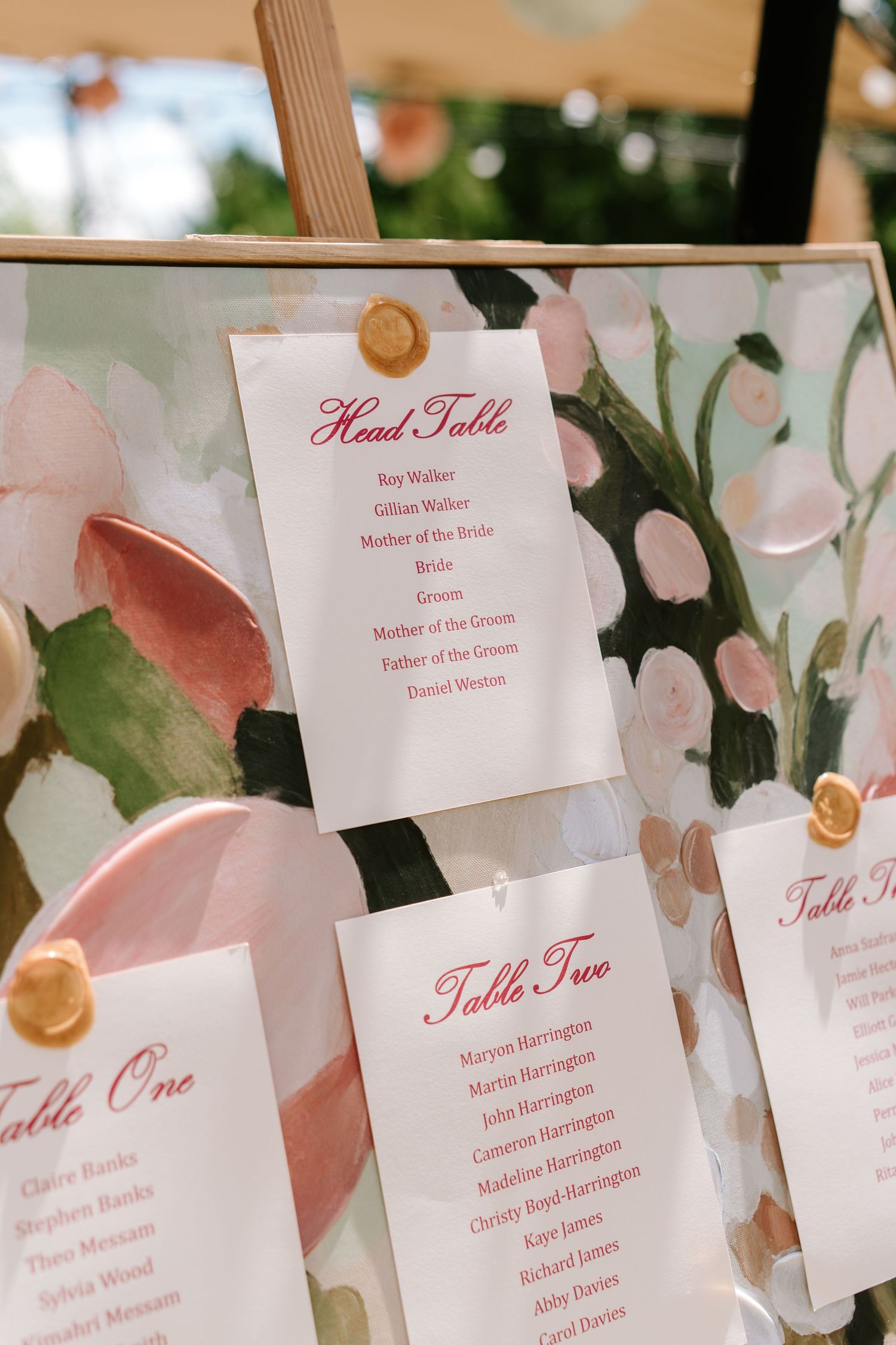 Elegant wedding seating chart displayed on a floral painted board with gold wax seal accents.