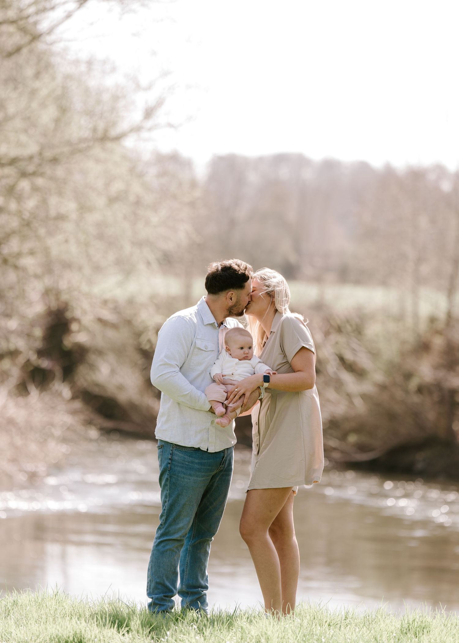 A couple shares a tender kiss over their newborn baby by a sunlit river in a serene outdoor family portrait.