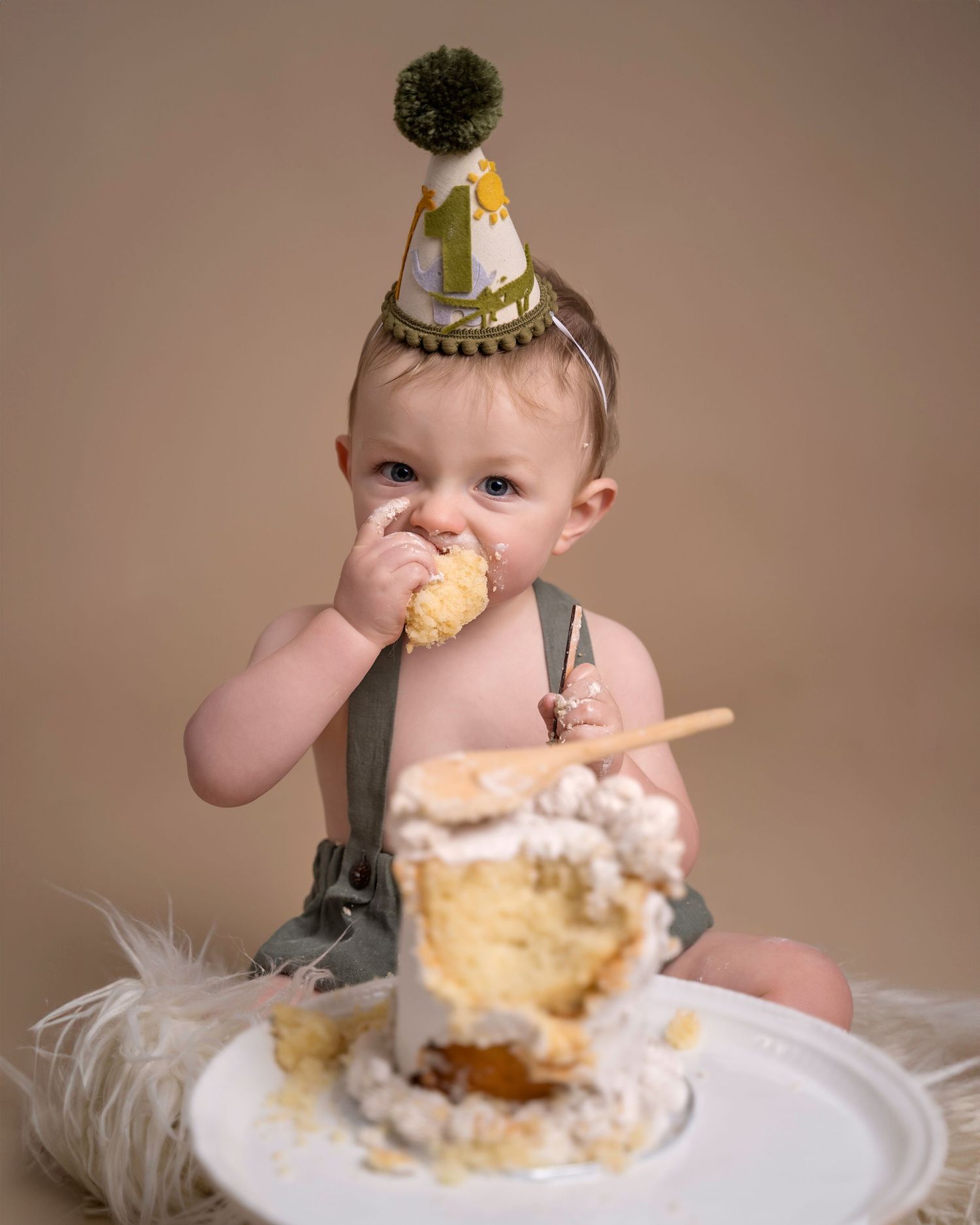 Baby in a party hat joyfully eating cake during a first birthday cake smash photo session.