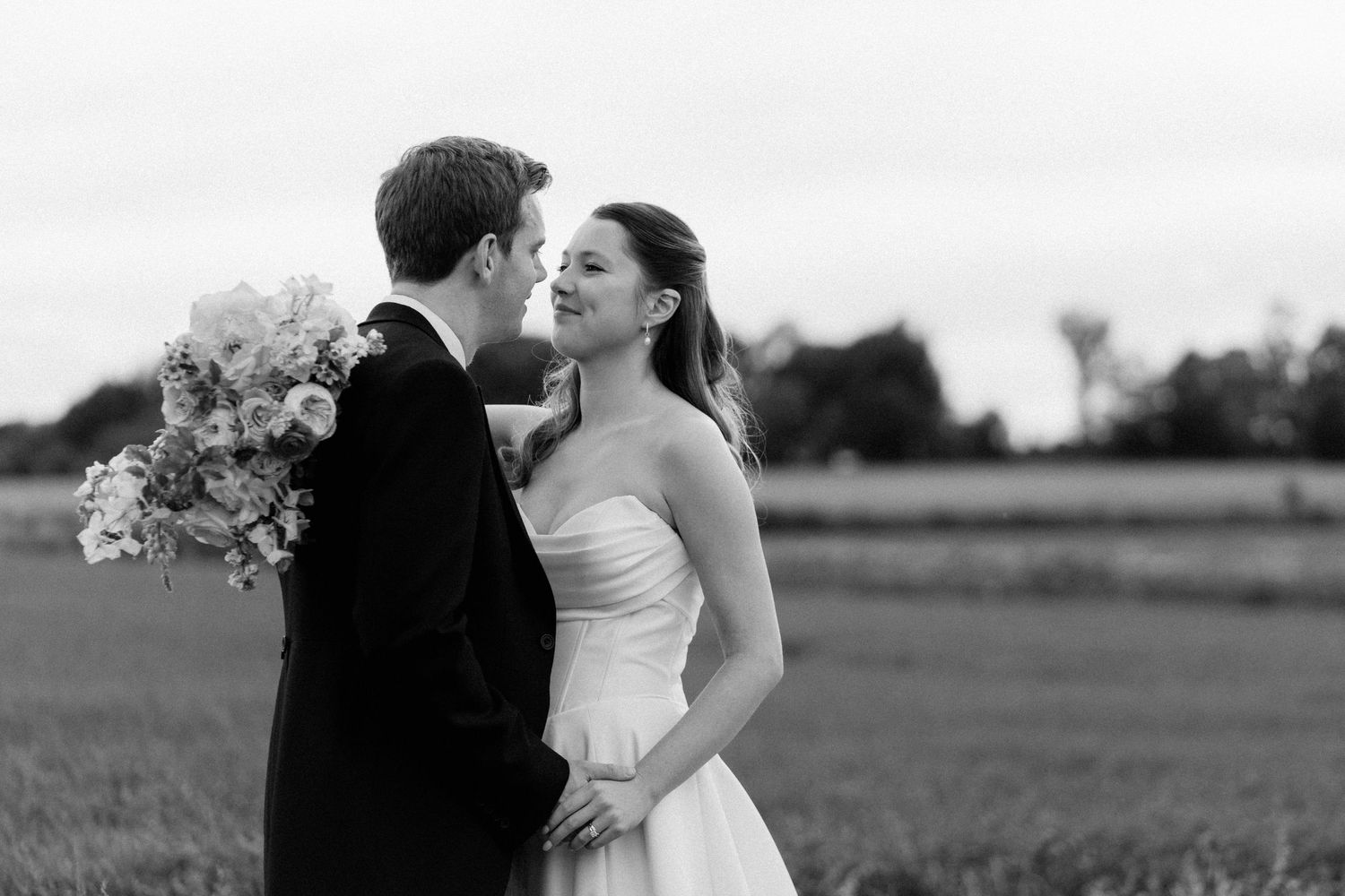 Black and white photo of a bride and groom sharing a romantic moment in a field, bride holding a bouquet.