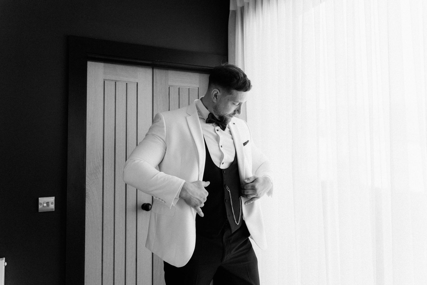 Black and white photo of a well-dressed groom adjusting his white blazer and bow tie before his wedding ceremony.