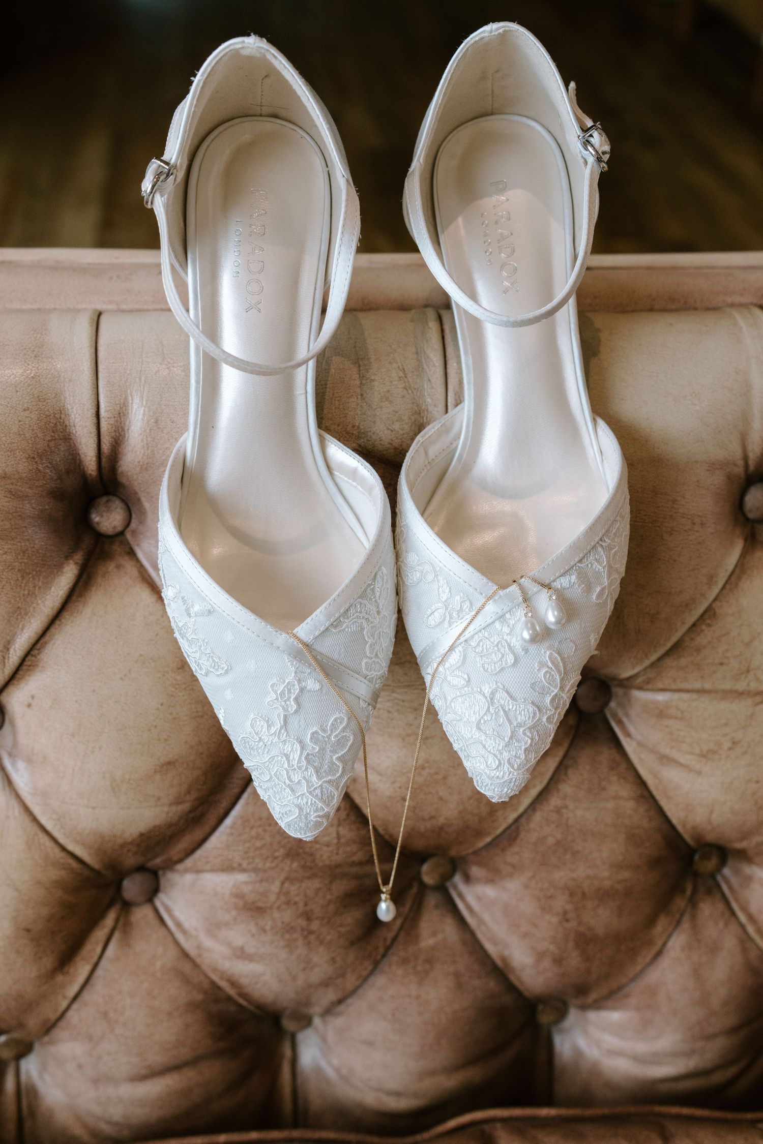 Elegant white lace bridal heels with ankle straps and pearl detail displayed on tufted beige leather surface.