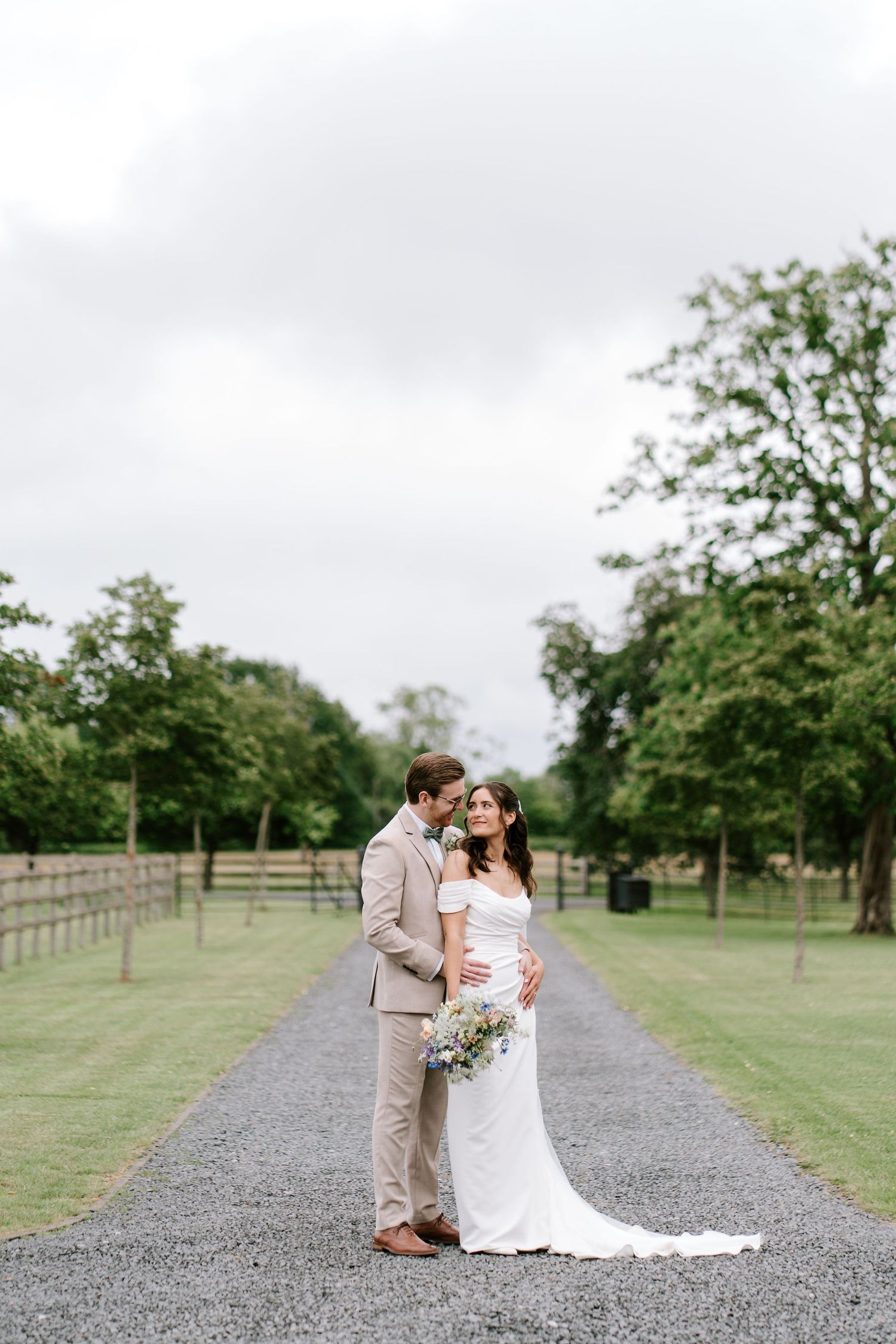Bride and groom embracing on a gravel path surrounded by lush green trees at an outdoor wedding venue.