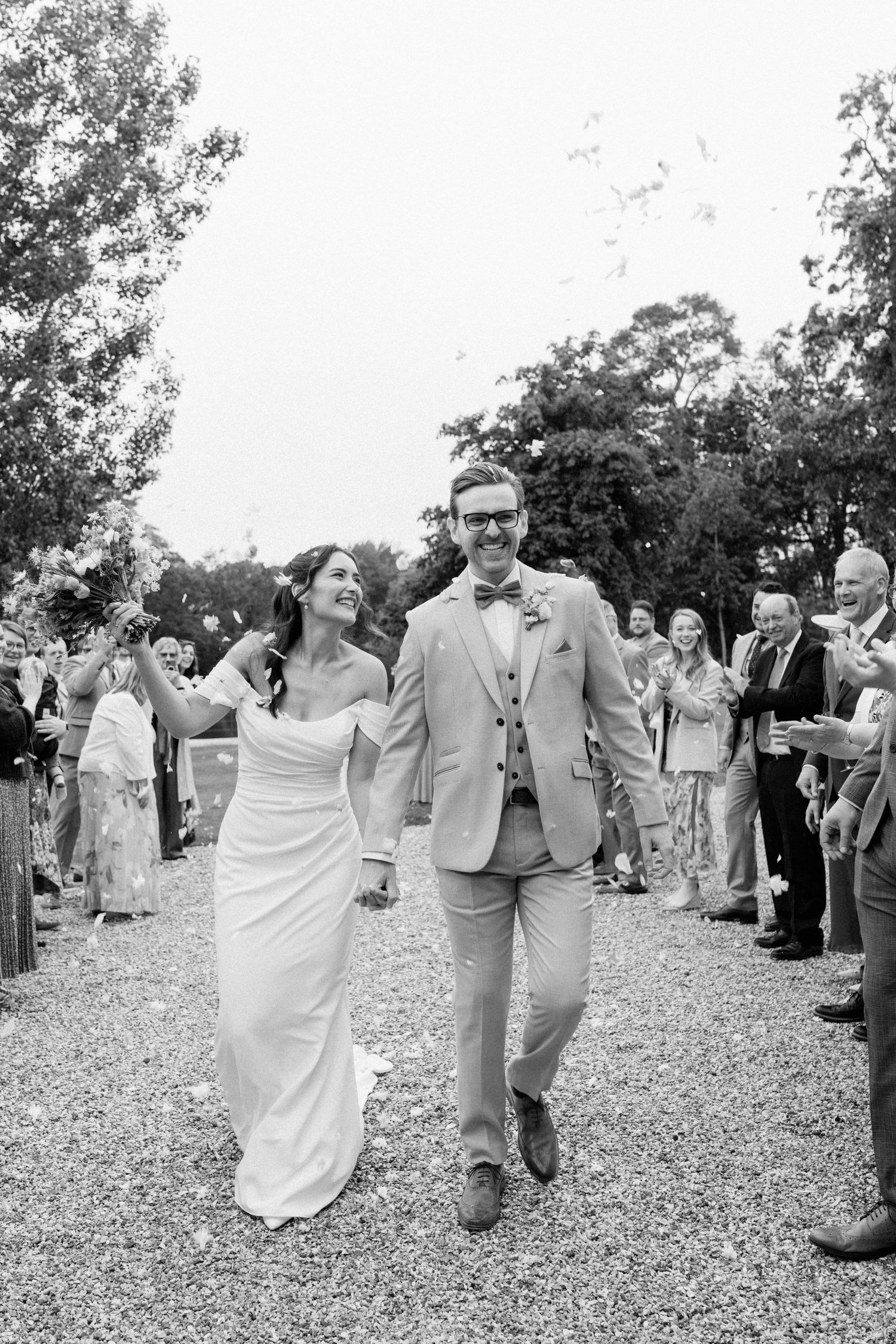 Black and white photo of smiling bride and groom walking through confetti lined aisle at outdoor wedding ceremony.