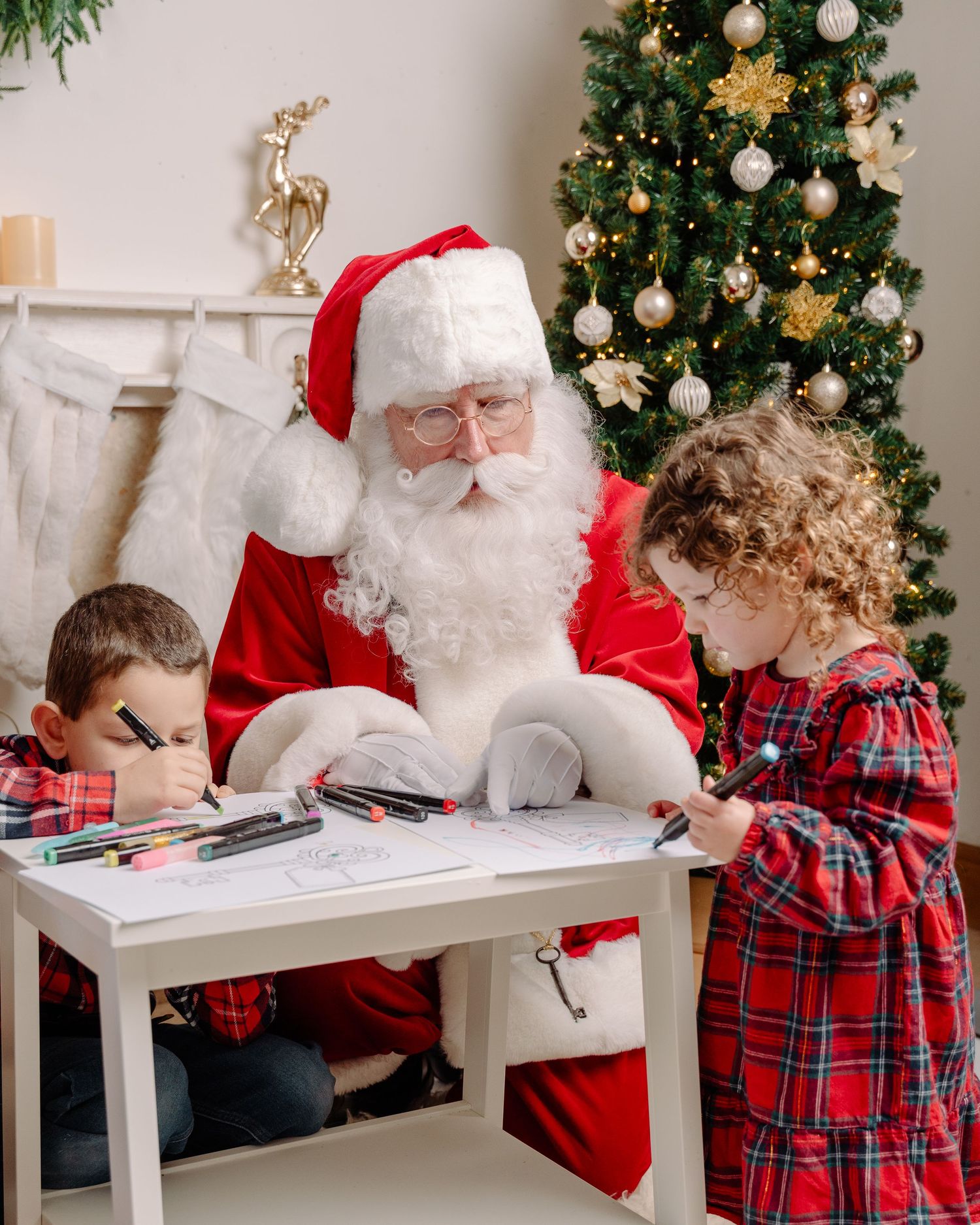 Santa Claus sits with two children in plaid outfits, drawing at a white table beside a decorated Christmas tree.