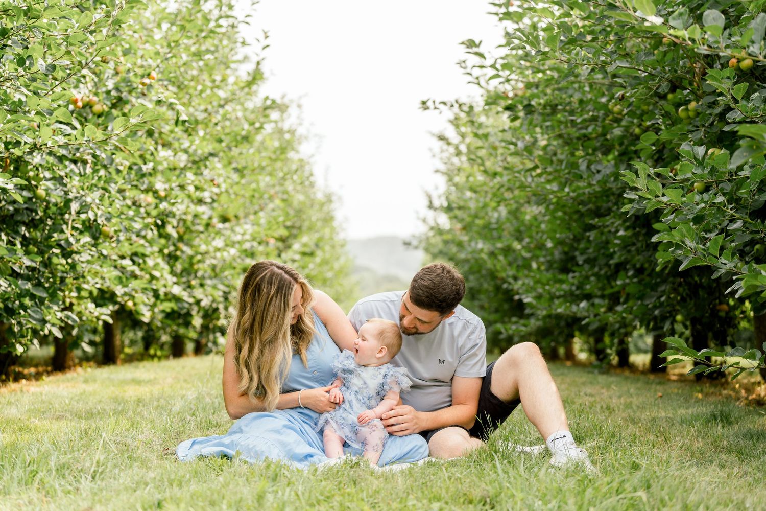 Family of three sitting on grass in an apple orchard, parents lovingly interacting with their baby girl.