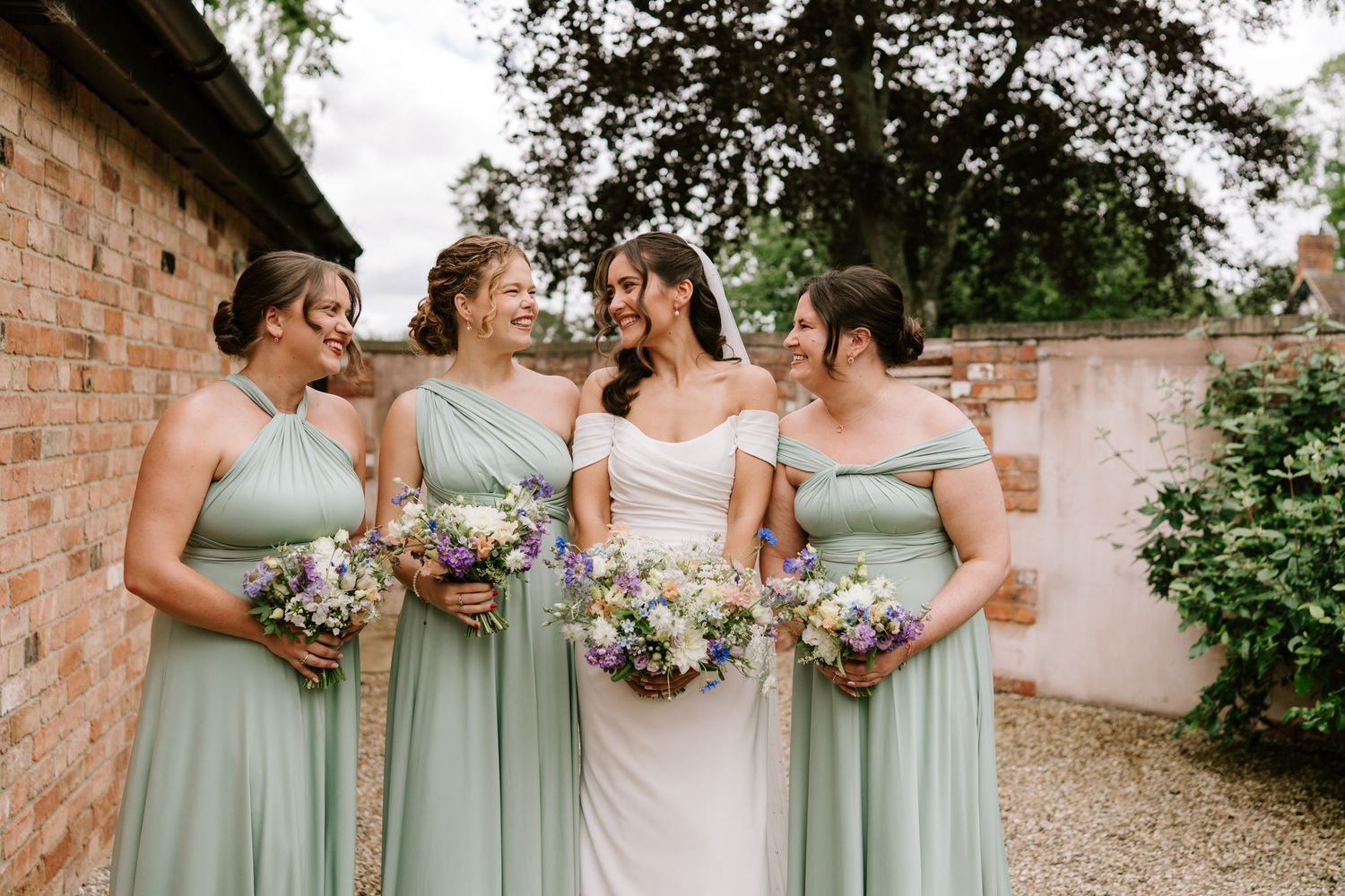 Bride in white gown with four bridesmaids in sage green dresses holding wildflower bouquets at an outdoor wedding venue.