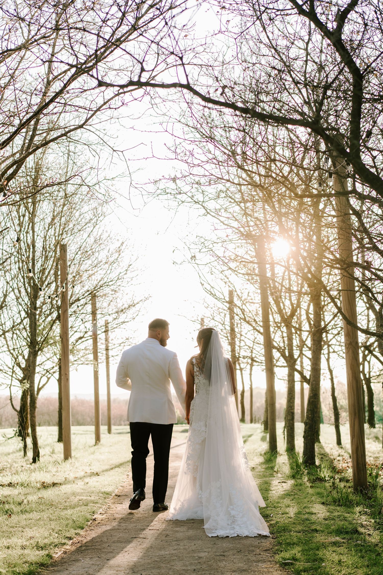 A bride and groom at Alrewas Hayes Country Estate walk hand in hand down a sunlit tree-lined path on their wedding day.