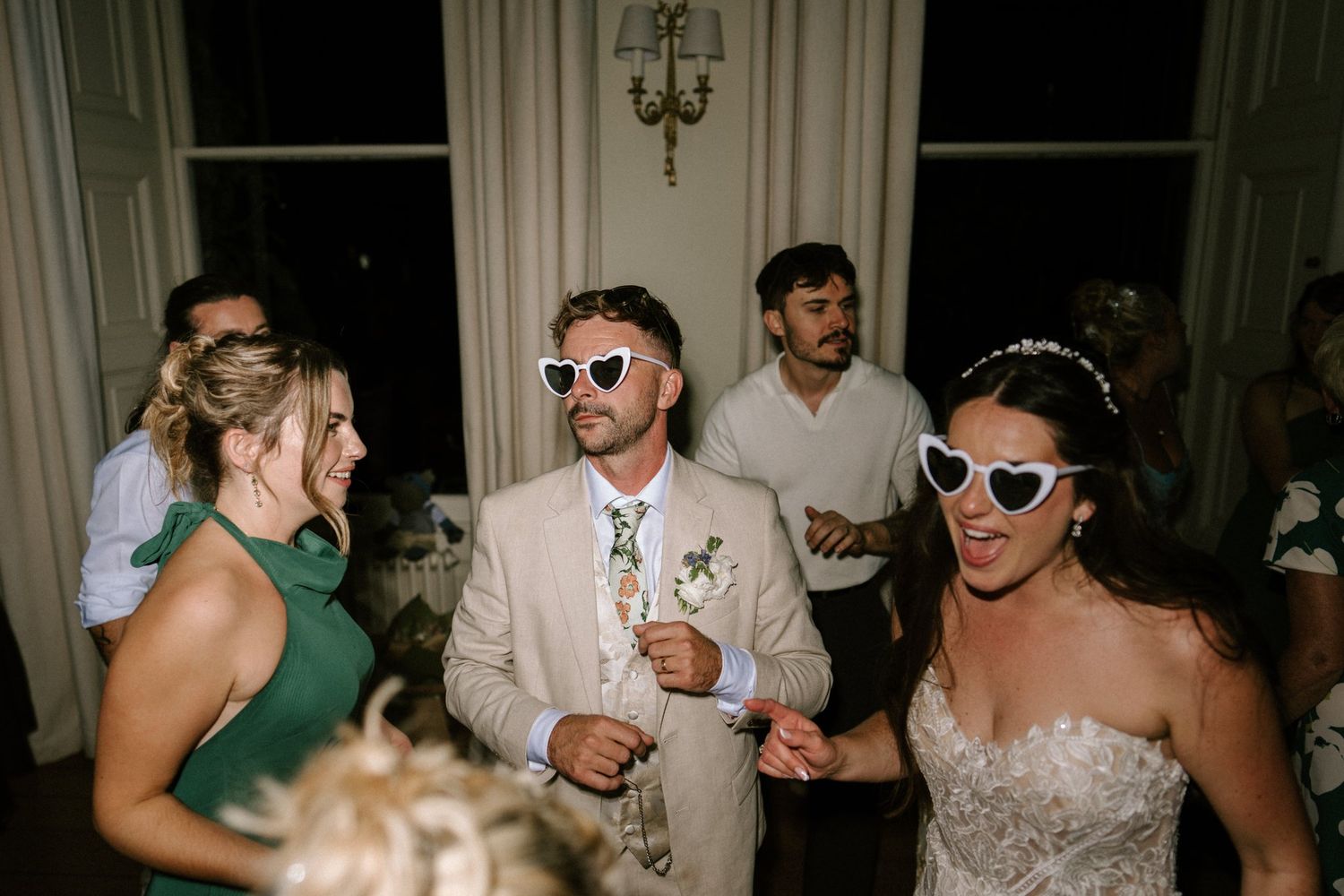 Bride and groom wearing white heart-shaped sunglasses dancing with guests at a wedding reception.