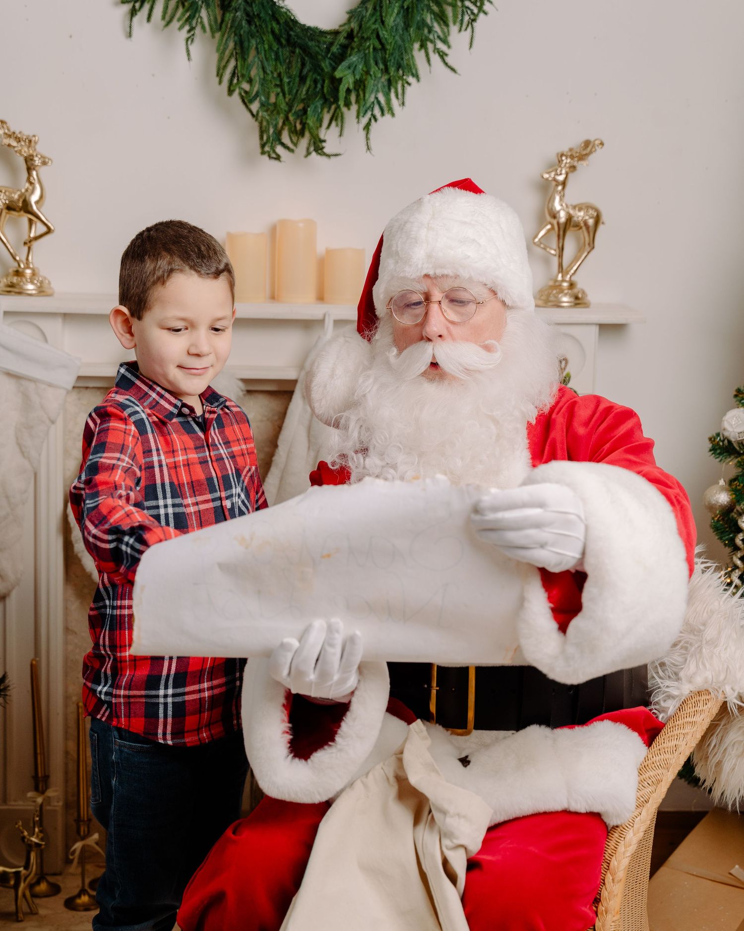 A young boy in plaid shirt stands beside Santa Claus reading a scroll in a Christmas decorated room.