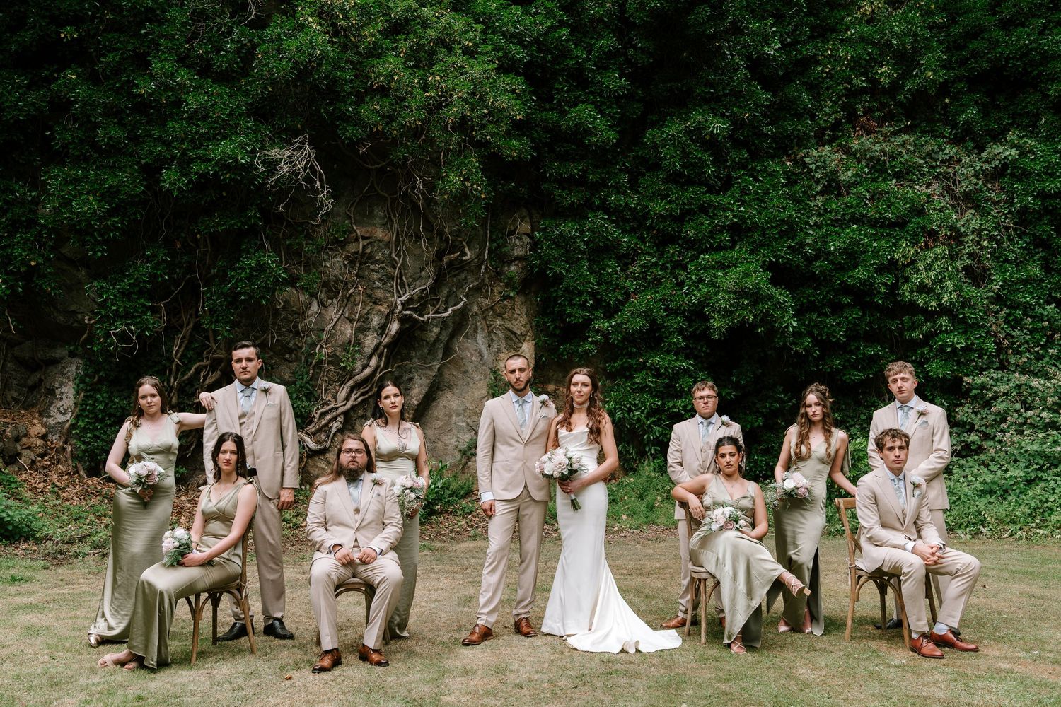 Wedding party in neutral tones posing outdoors with lush green cliffs and trees in the background.