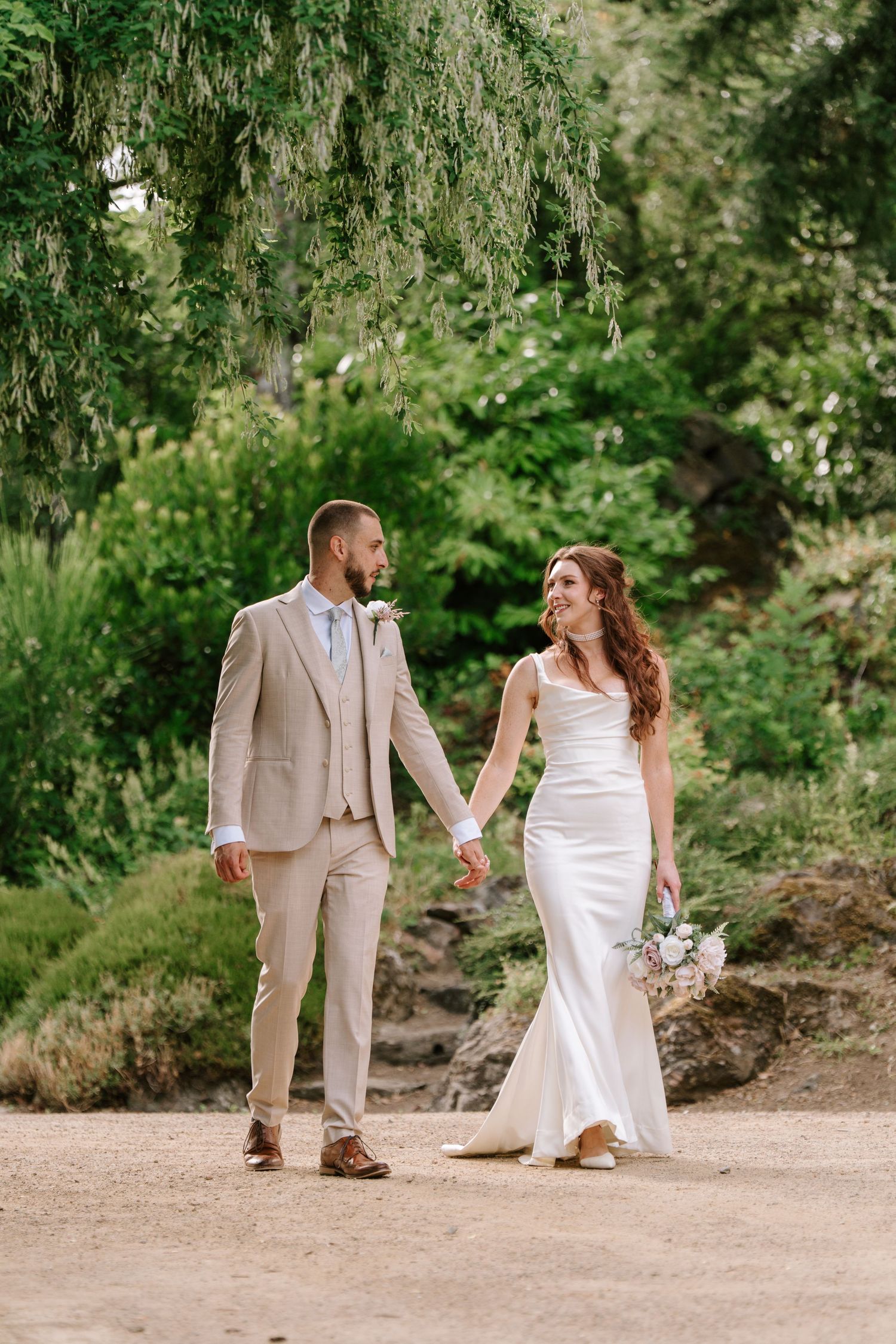 Bride and groom walk hand in hand through a lush green garden on their wedding day.
