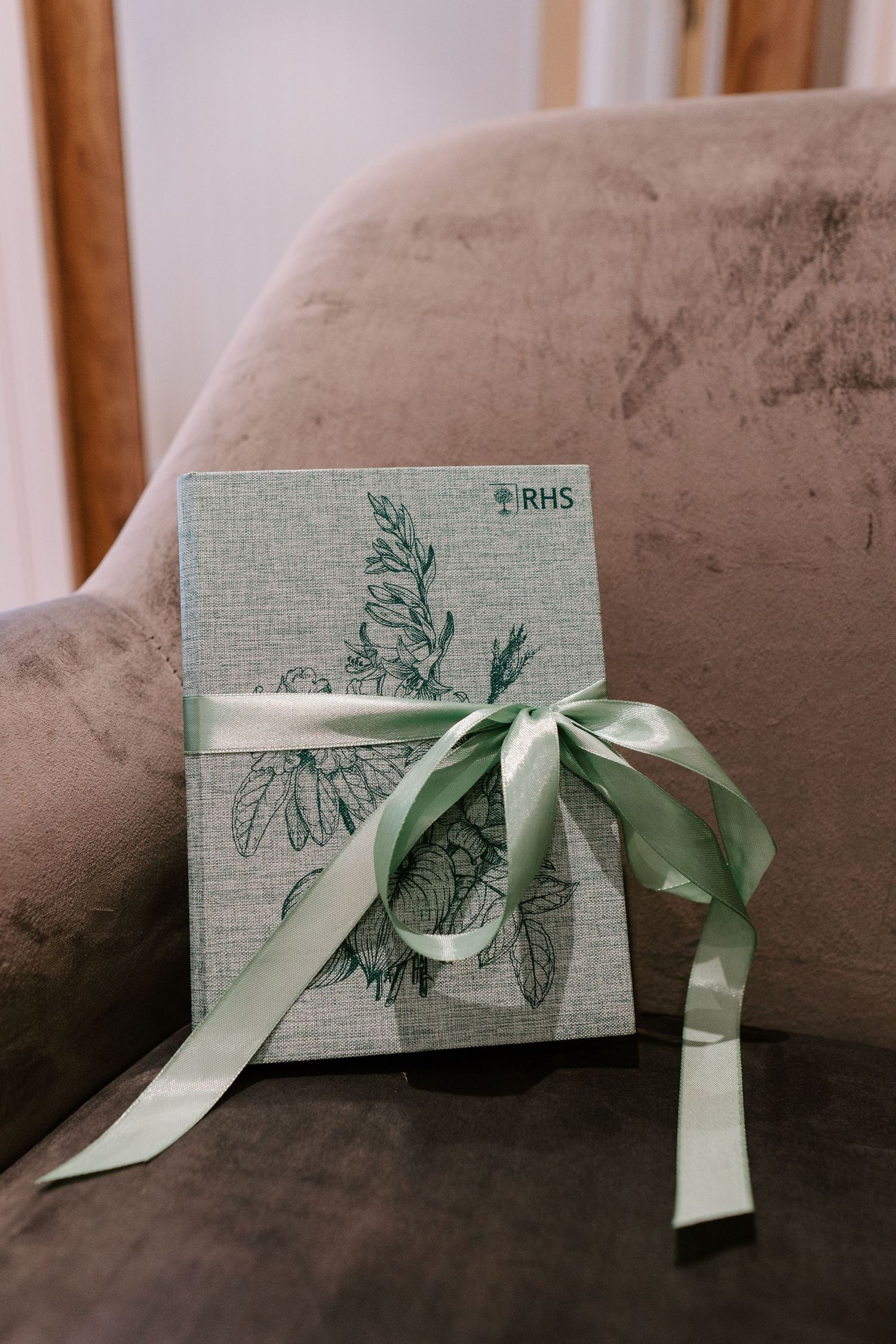 RHS botanical journal with green ribbon bow resting on a velvet chair at a wedding.
