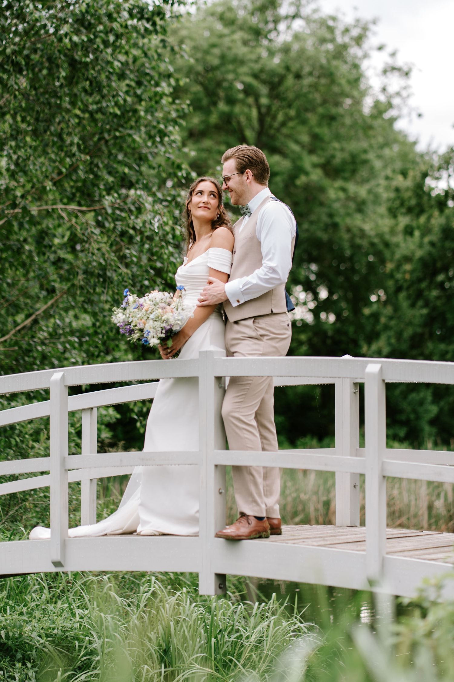 Bride and groom embracing on white arched bridge surrounded by lush green garden.
