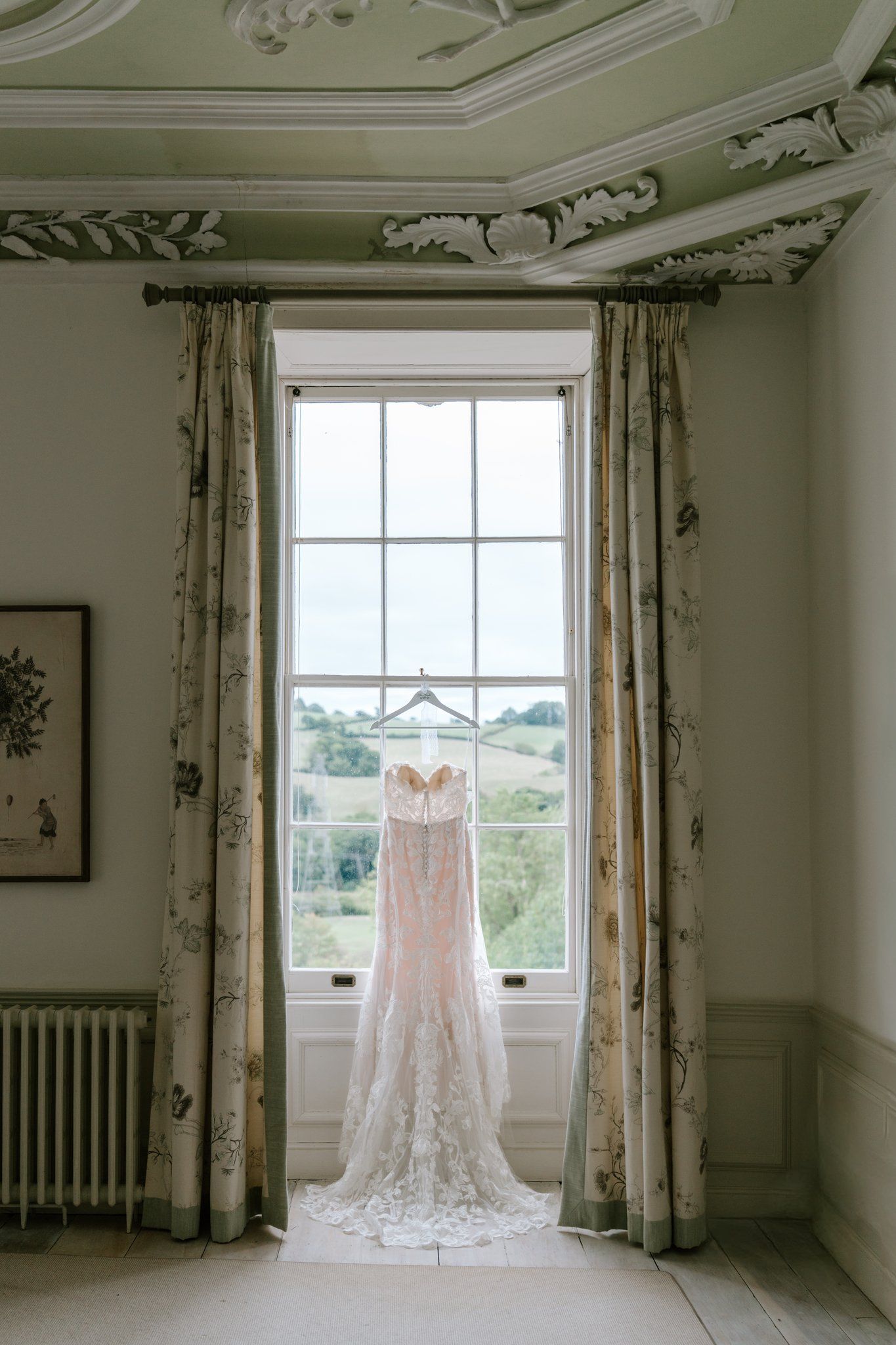 Elegant lace wedding dress hanging in a window with floral curtains and ornate green ceiling molding in a stately home.
