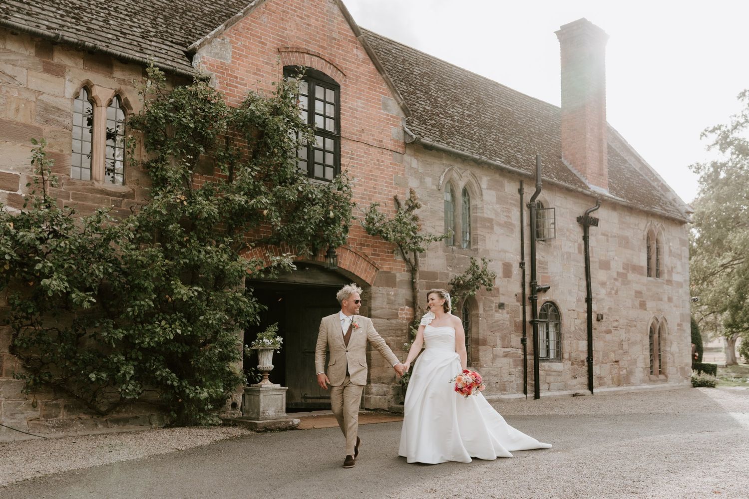 Bride and groom walking hand in hand past the sandstone manor house at Brinsop Court, Herefordshire