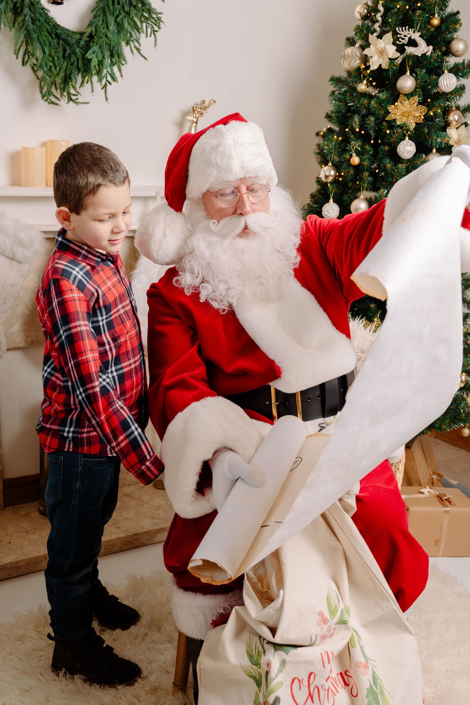 A young boy stands beside Santa Claus reading a scroll near a decorated Christmas tree indoors.