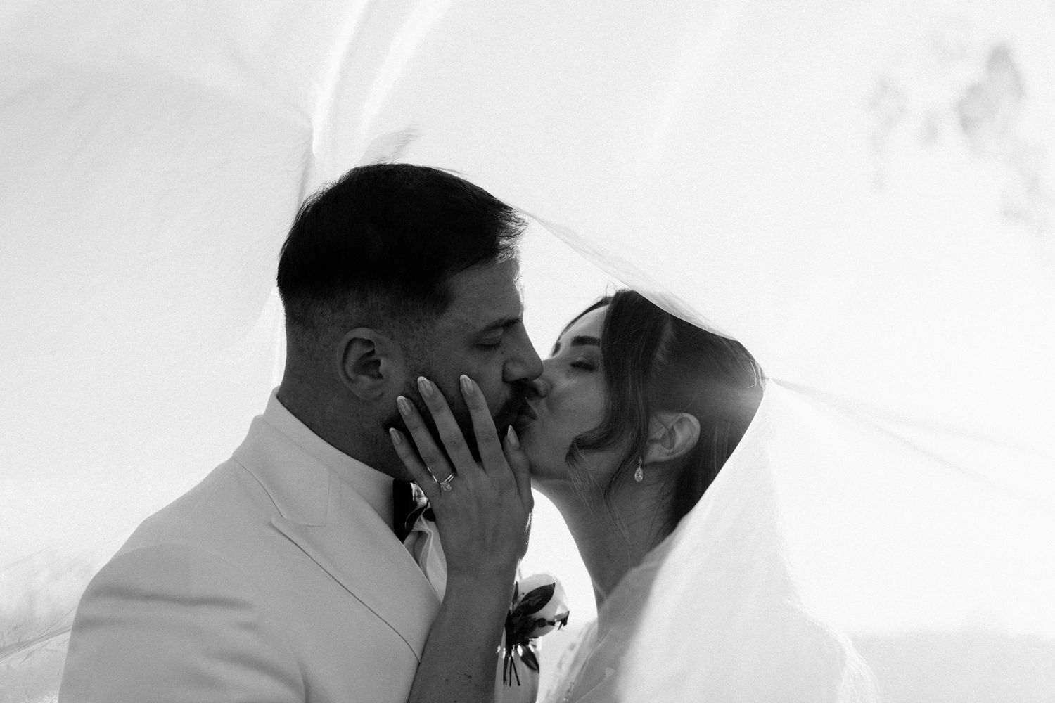 Black and white photo of a bride and groom sharing a romantic kiss under a flowing wedding veil.