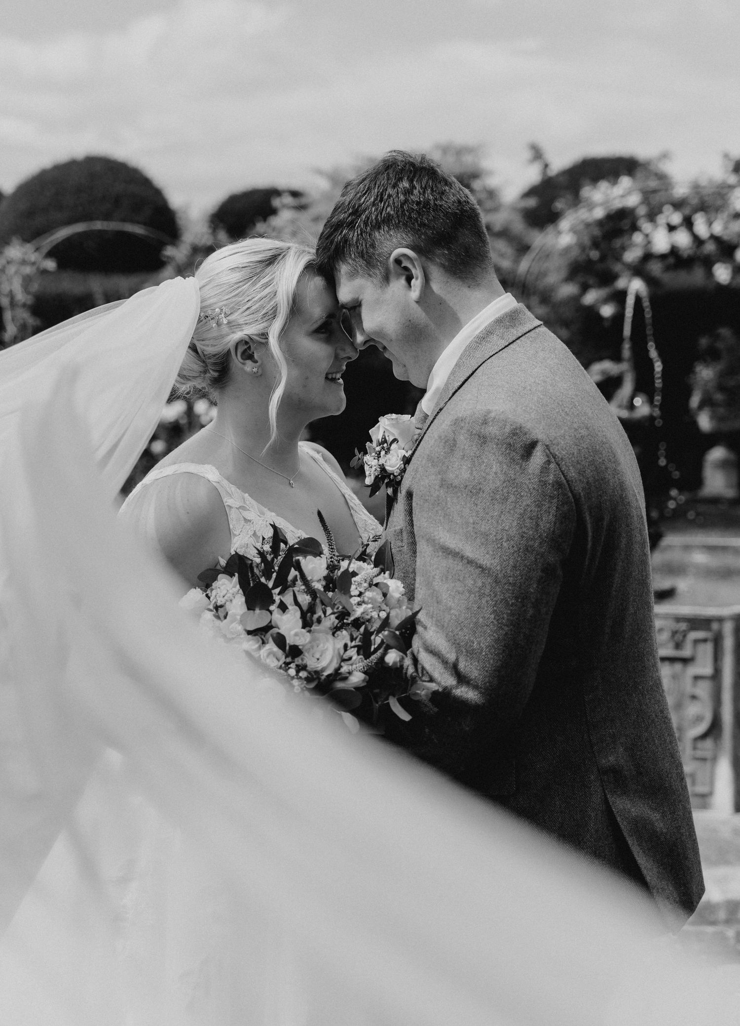 Black and white wedding photo of bride and groom touching foreheads, veil flowing in wind, holding bouquet outdoors.
