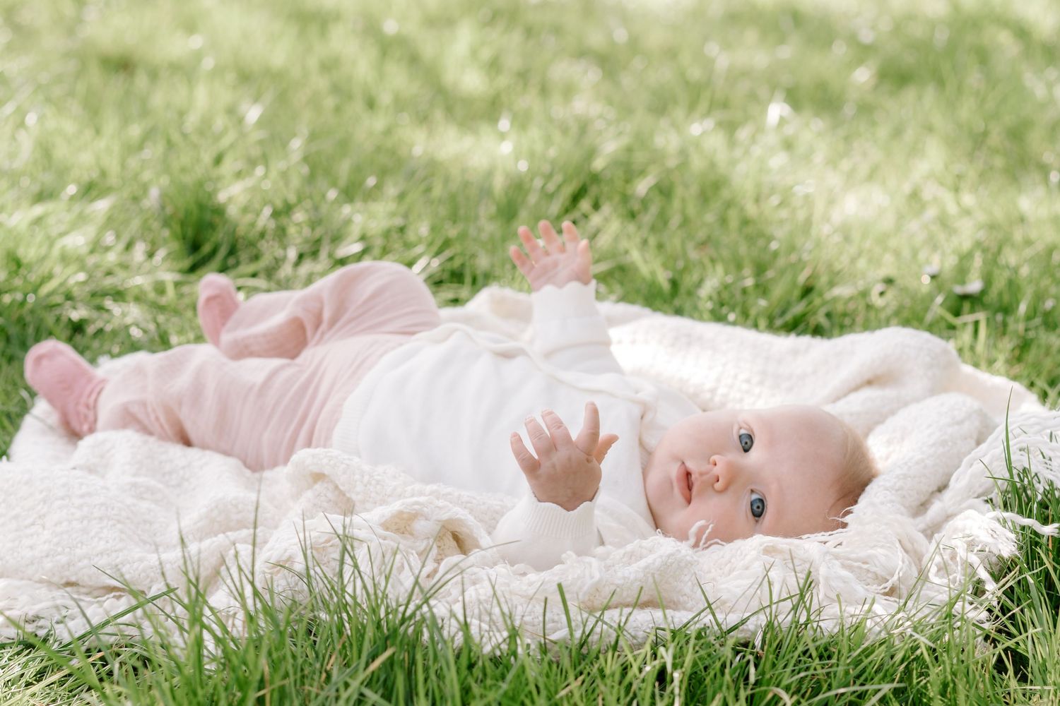 A happy baby in pink outfit lying on a white blanket in lush green grass outdoors on a sunny day.