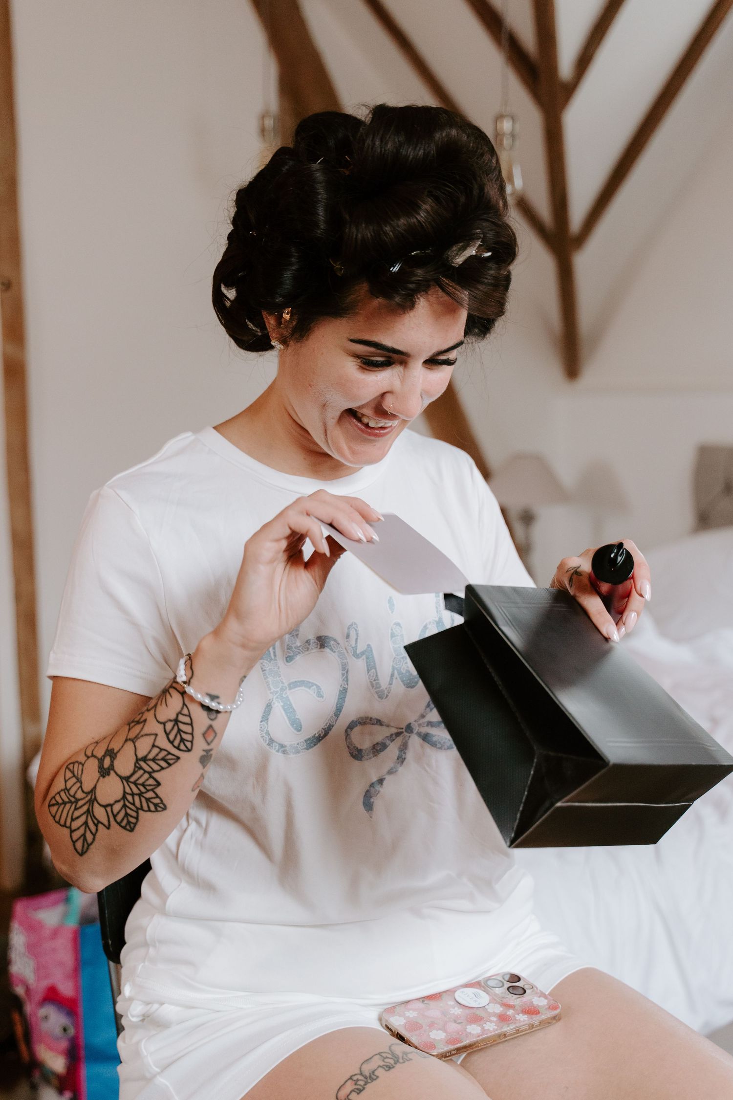 Young woman with hair rollers and tattoos smiling while opening a black gift bag on a bed.