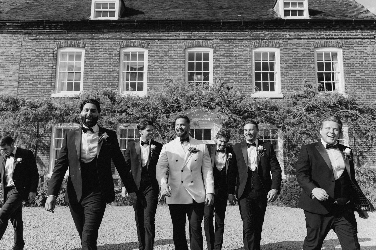 Black and white photo of a groom in white tuxedo walking with groomsmen in black suits outside an elegant brick manor.