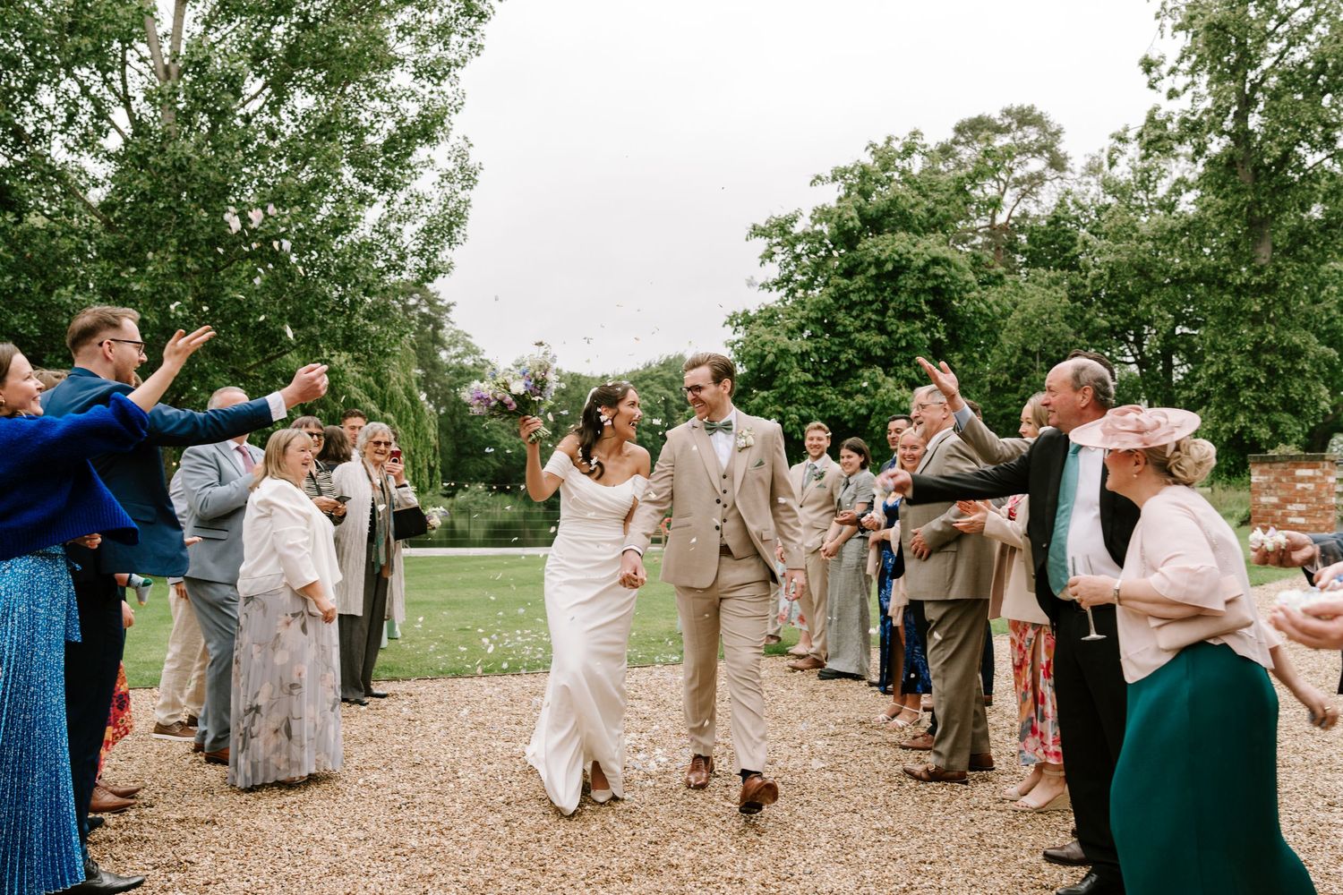 Bride and groom walk through confetti shower surrounded by cheering wedding guests outdoors.