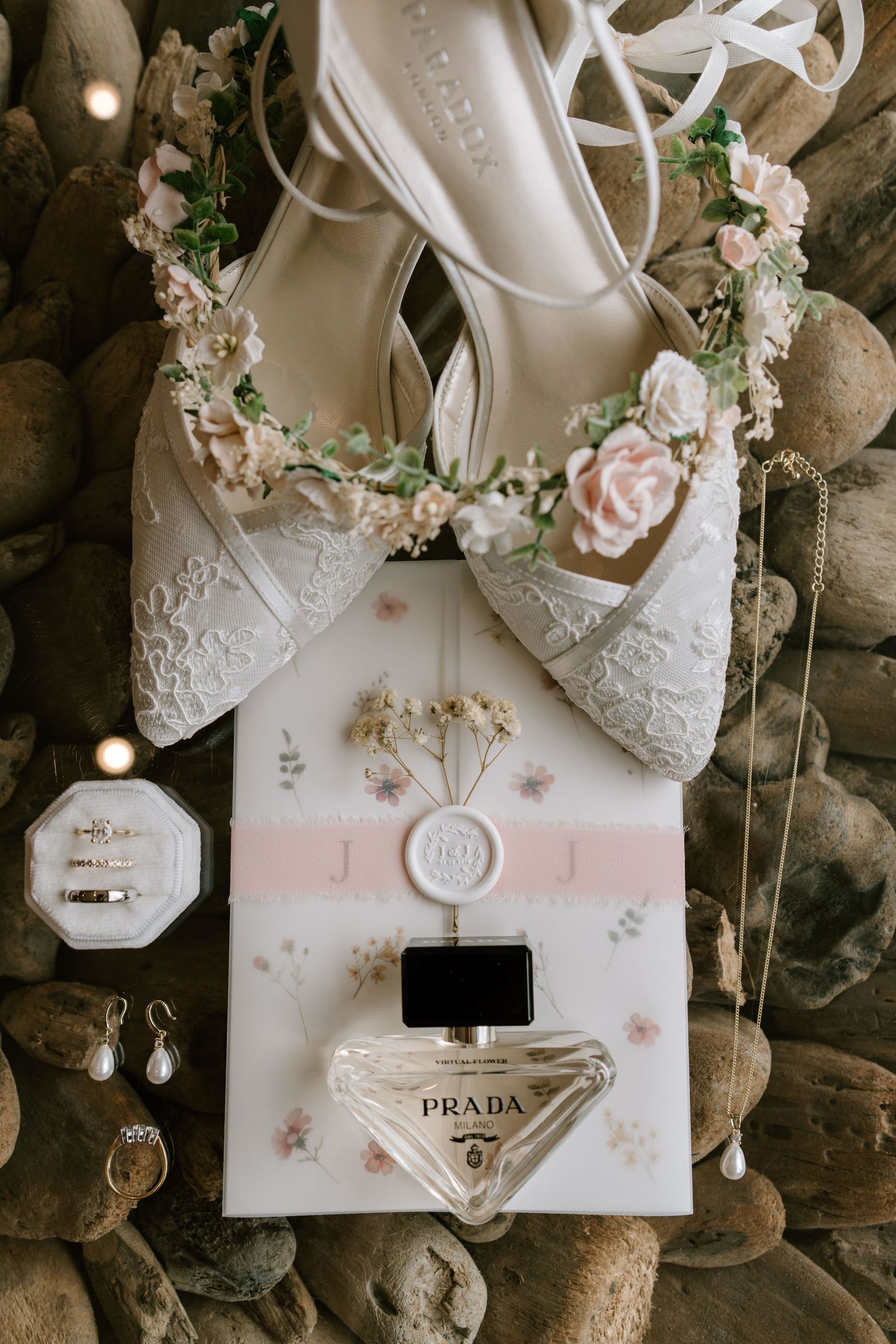 Elegant bridal flat lay featuring white lace floral shoes, Prada perfume, gold rings, pearl earrings, and a monogram wax seal card.