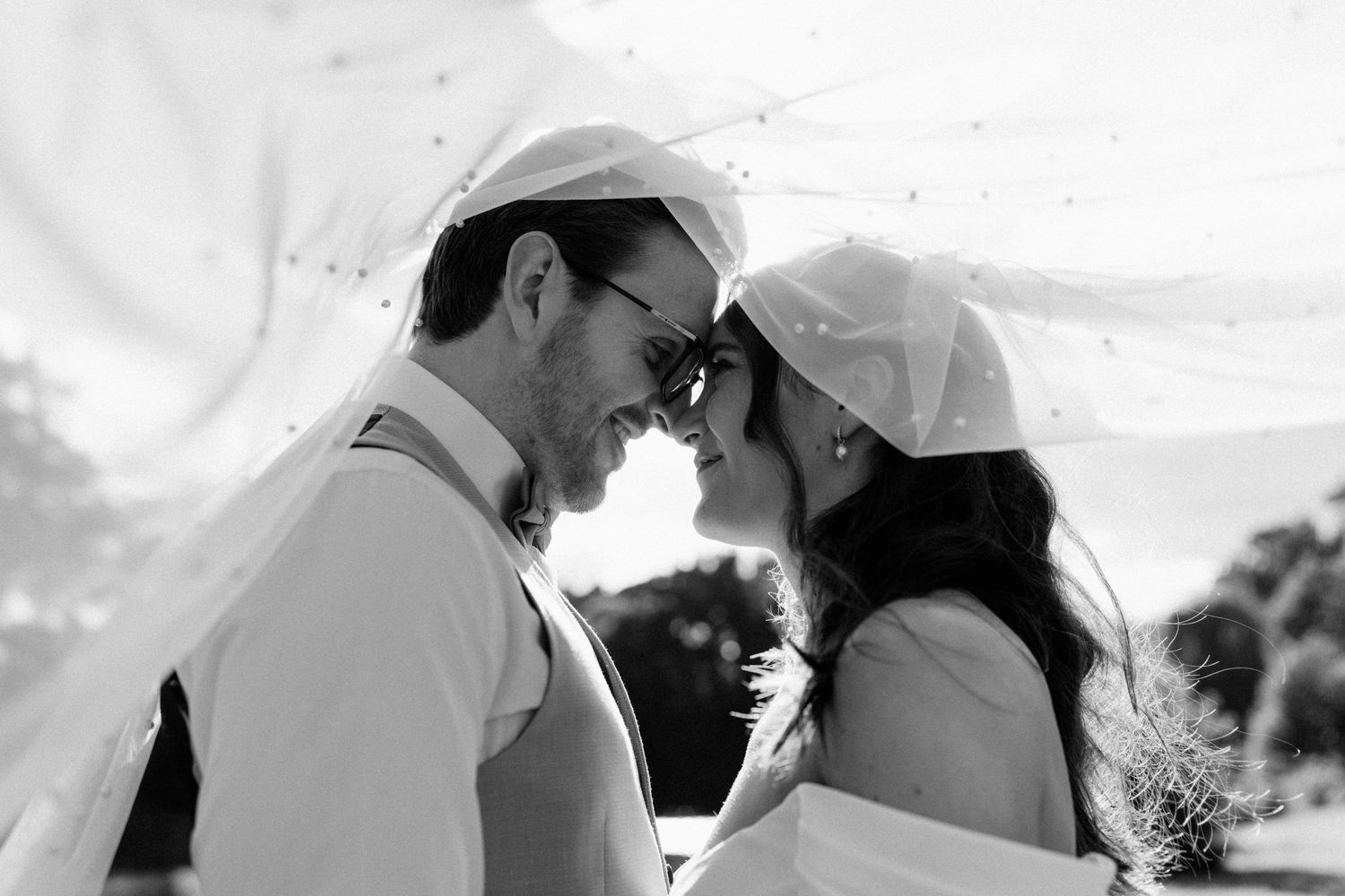 Black and white photo of a bride and groom smiling under a flowing veil on their wedding day.