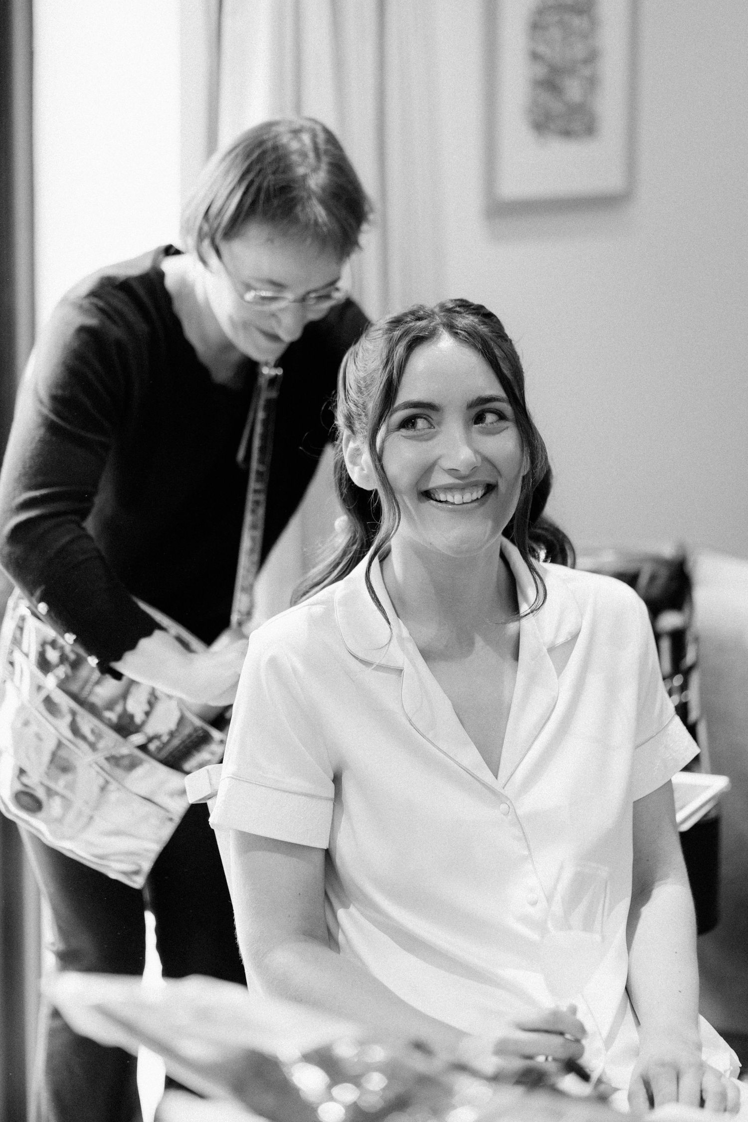 Bride smiling during wedding preparations while hairstylist works in background, black and white photo.