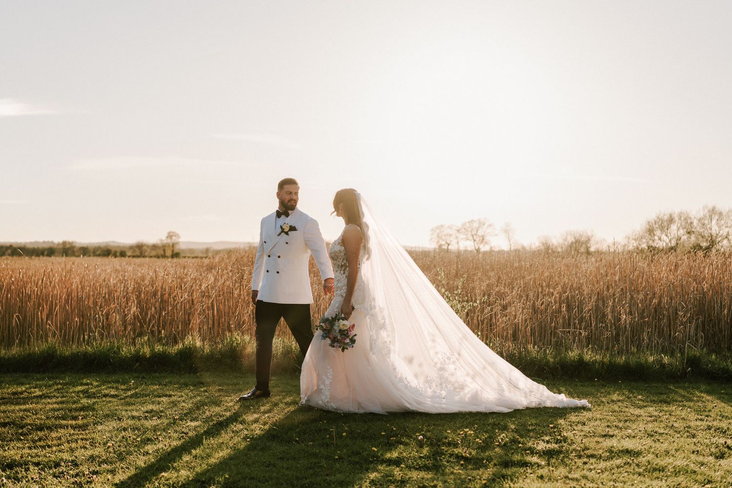 Bride and groom holding hands in golden field at sunset, bride in flowing white gown with long train.
