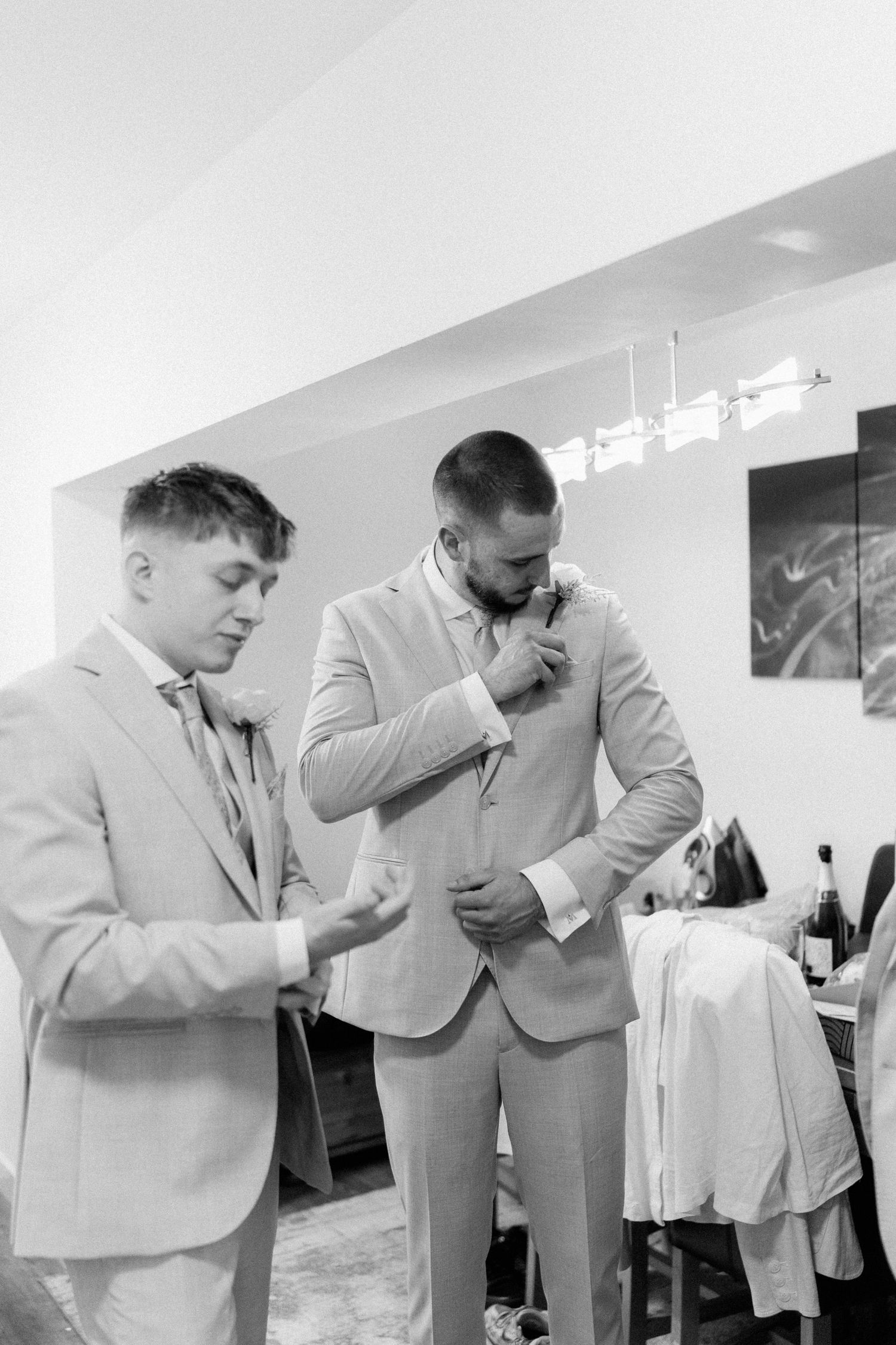 Two groomsmen in light suits help each other with boutonnieres while getting ready for a wedding.