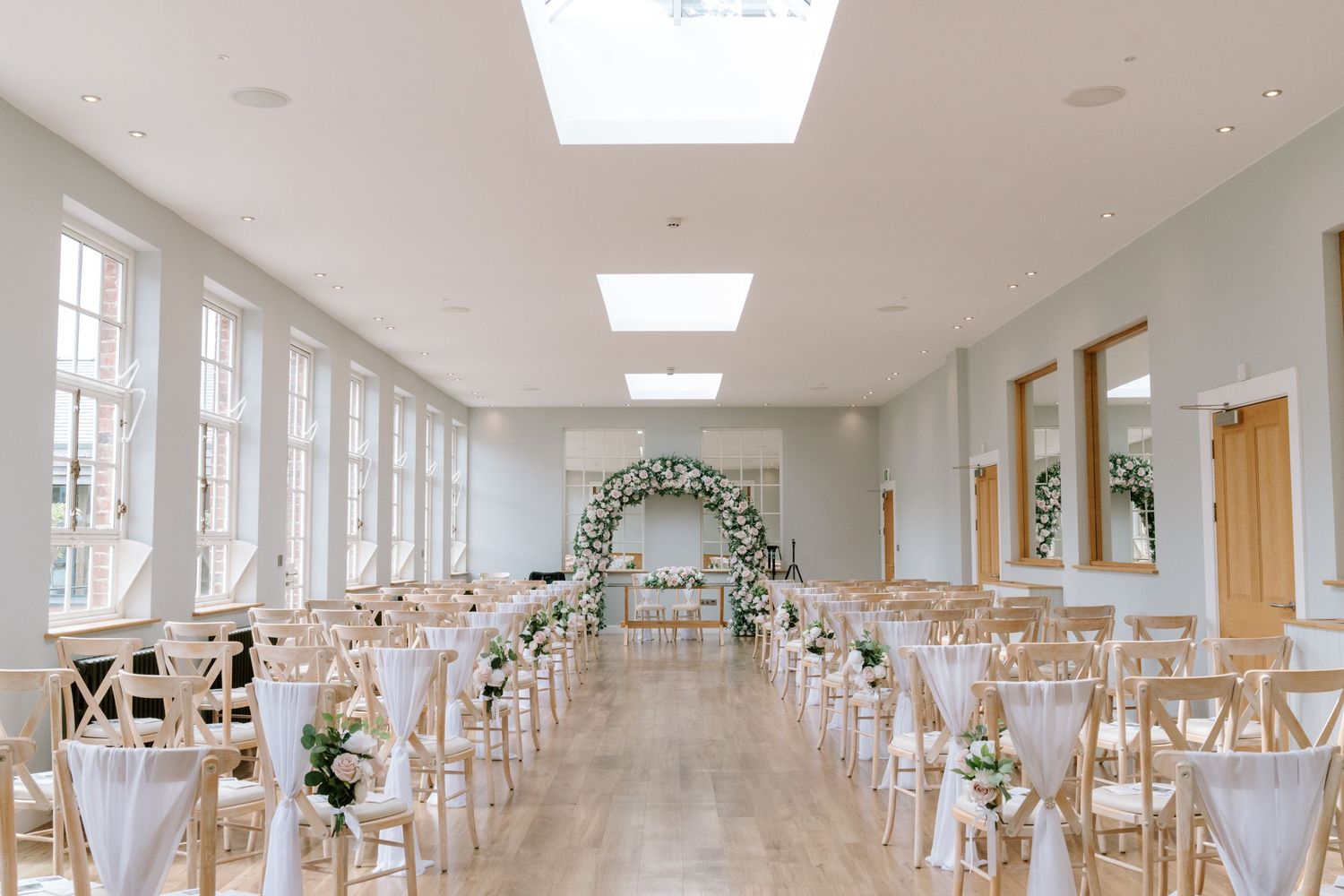 Elegant wedding ceremony venue with white chairs, floral arch, skylights, and soft natural light in a bright, airy room.