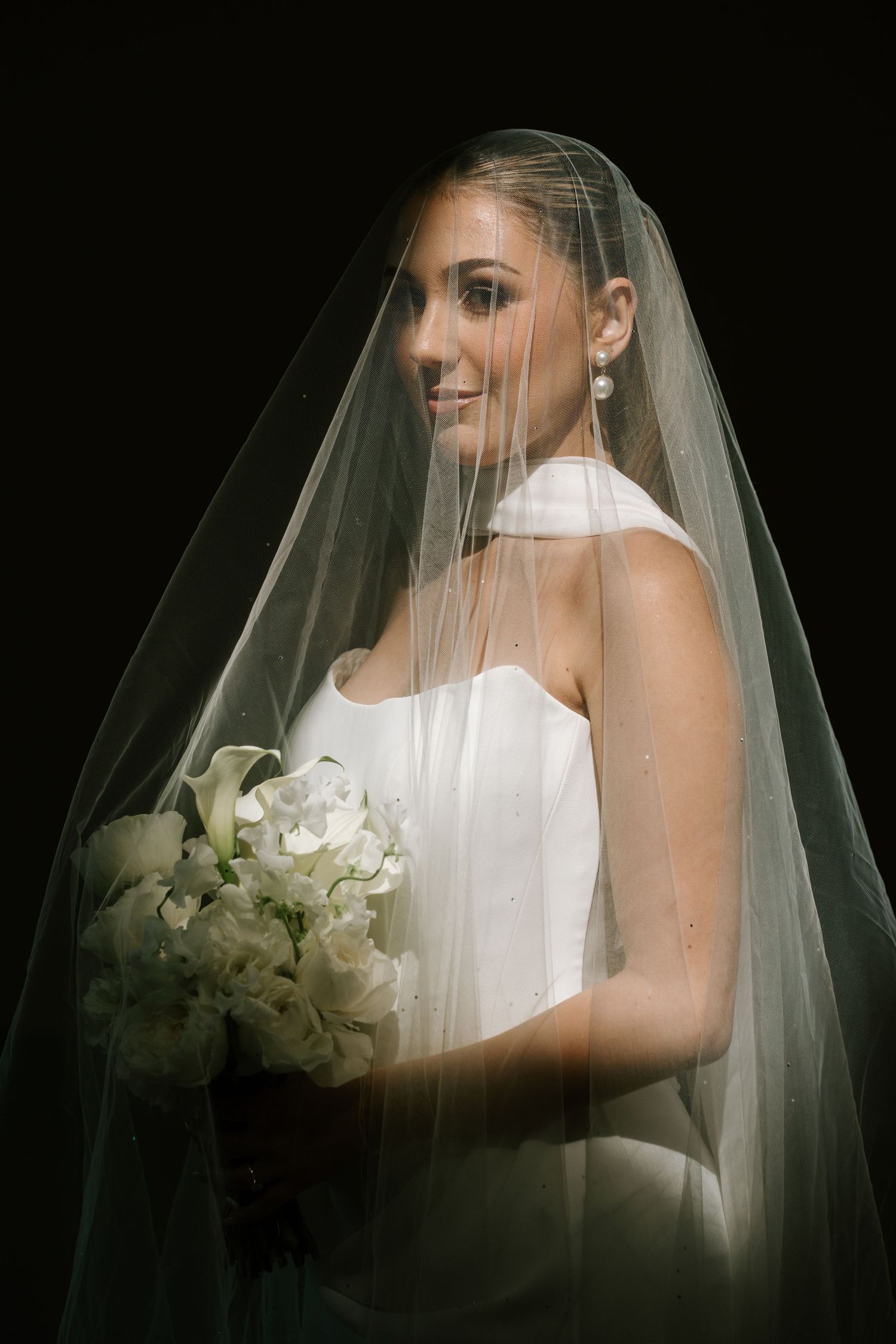 Elegant bride in white strapless gown and veil holding white floral bouquet, smiling against dark background.