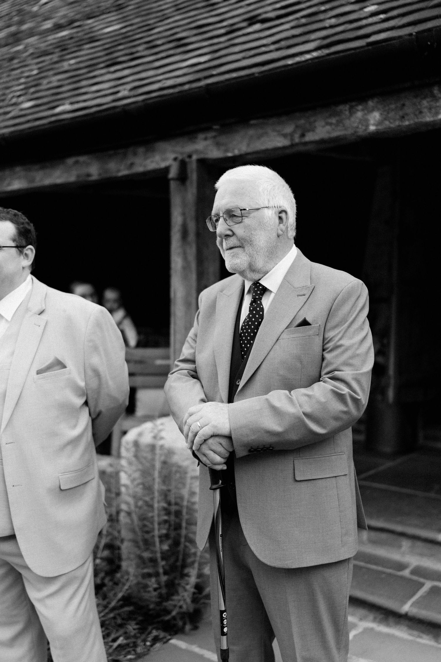 Elderly gentleman in a suit leaning on a cane at an outdoor wedding venue in black and white photo.