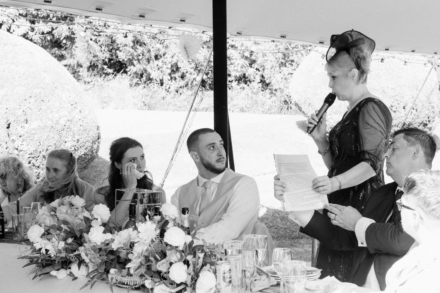 Black and white photo of woman giving speech at outdoor wedding reception with floral centerpieces.