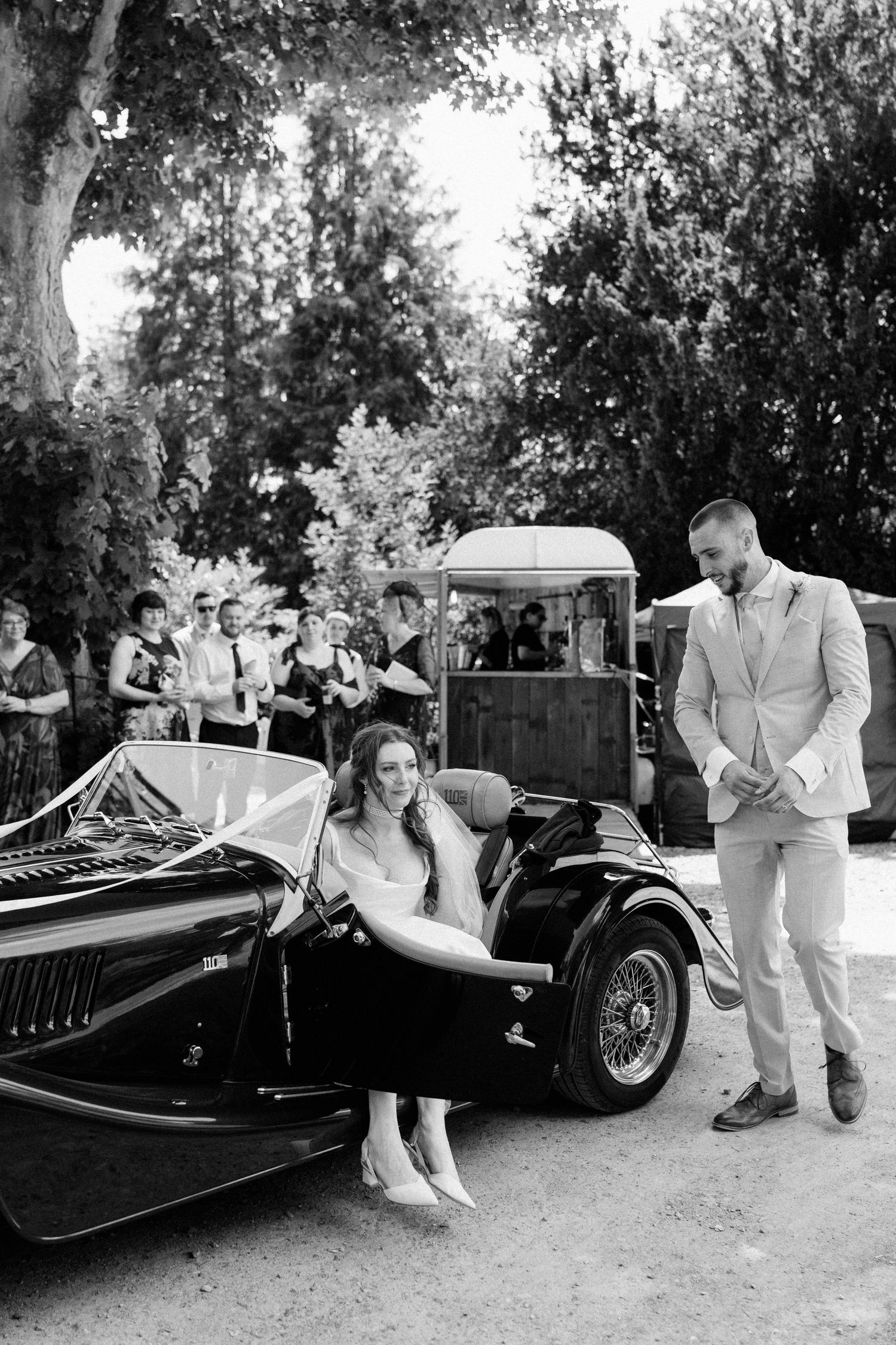 Bride and groom posing with vintage black convertible car at outdoor wedding in black and white photo.