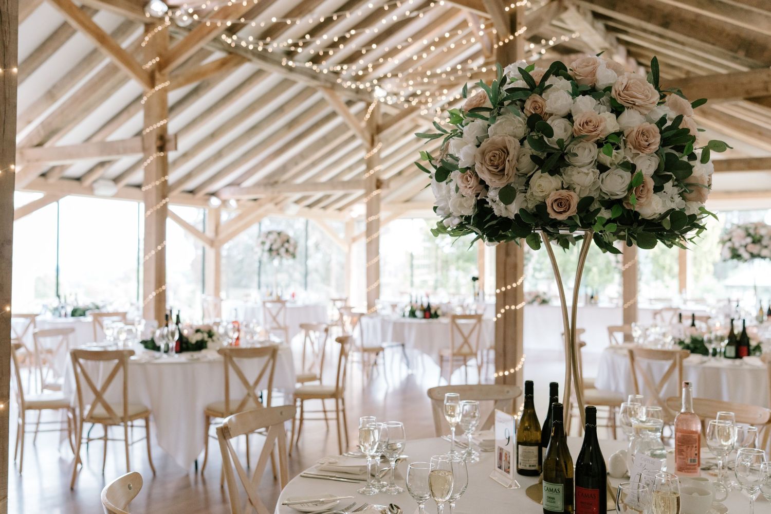 Elegant wedding reception venue with floral centerpieces, fairy lights, white linens, and wine bottles on round tables.